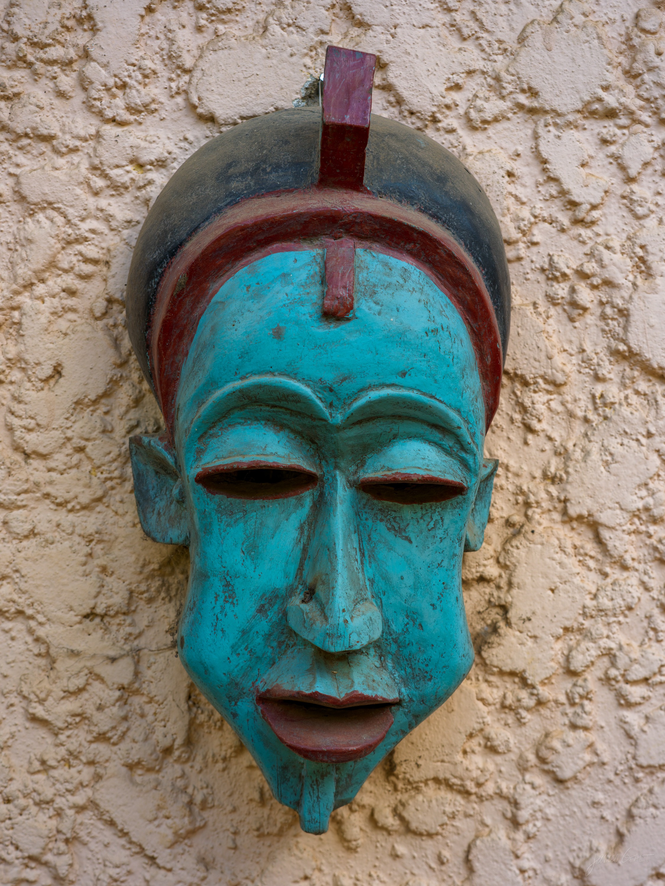 A carved wooden mask painted matte blue with a red crest and pale eyes, hung on a rough cream stucco wall.
