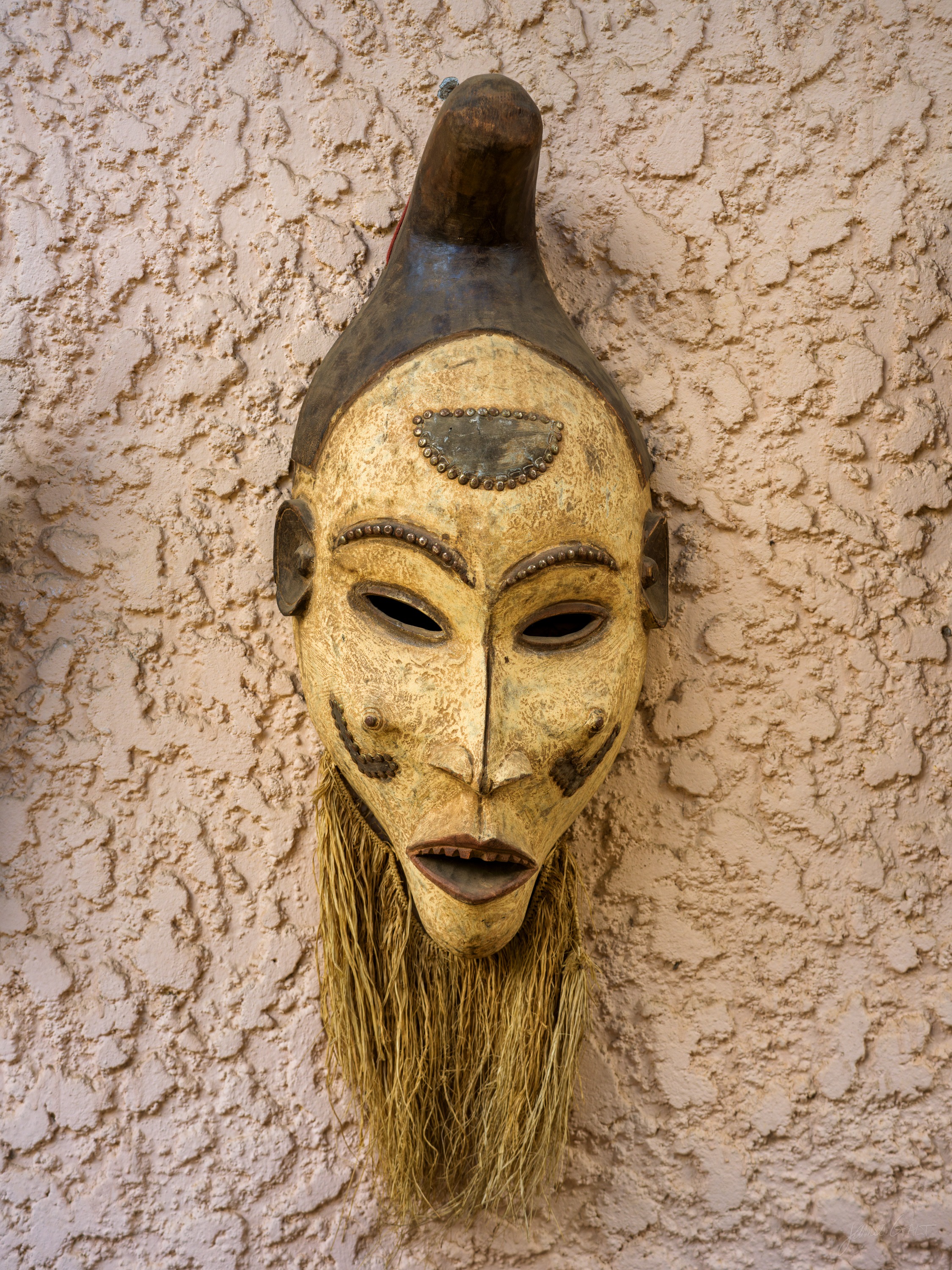 A tall, narrow carved mask with an elongated face and a fibre beard, mounted on a textured cream wall.