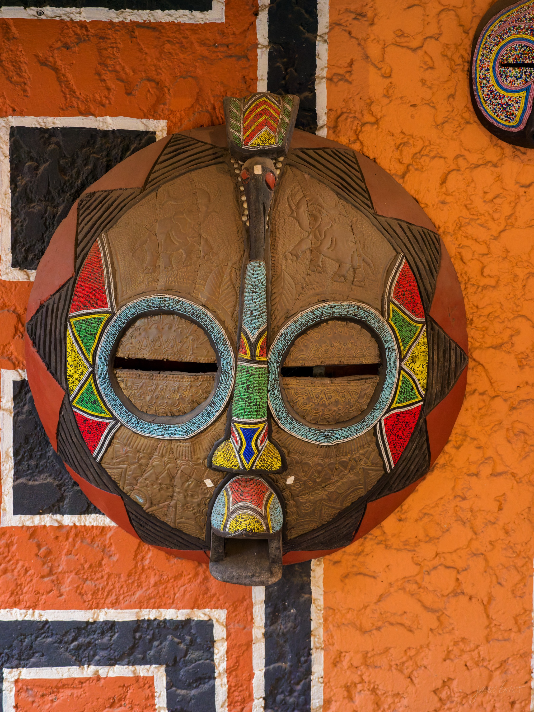 A round mask with a bold painted face in red, blue and cream, mounted on a bright orange wall.