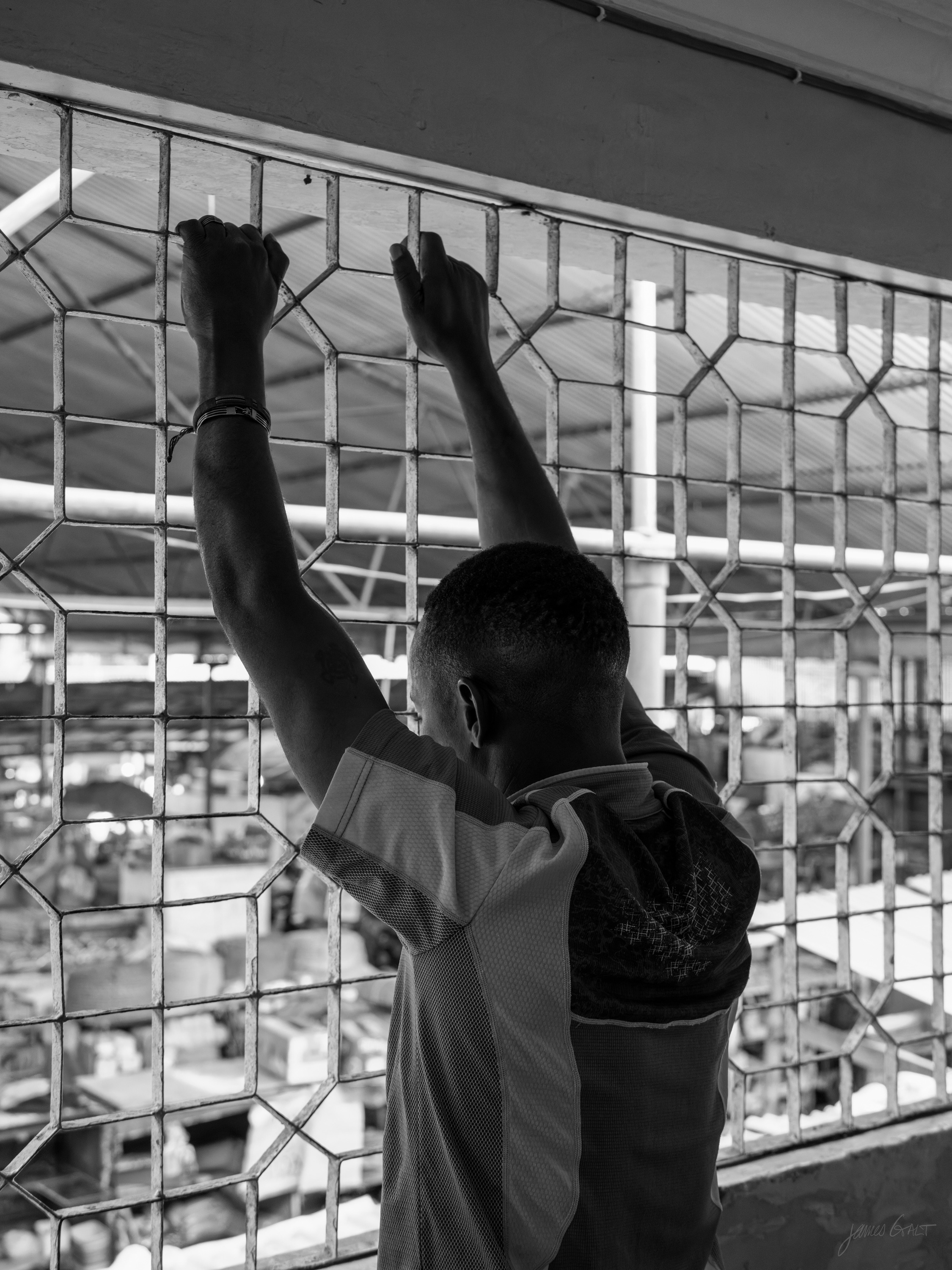 Black and white photograph of a young man seen from behind, both arms raised to the metal grille of a market stall, a busy market beyond.