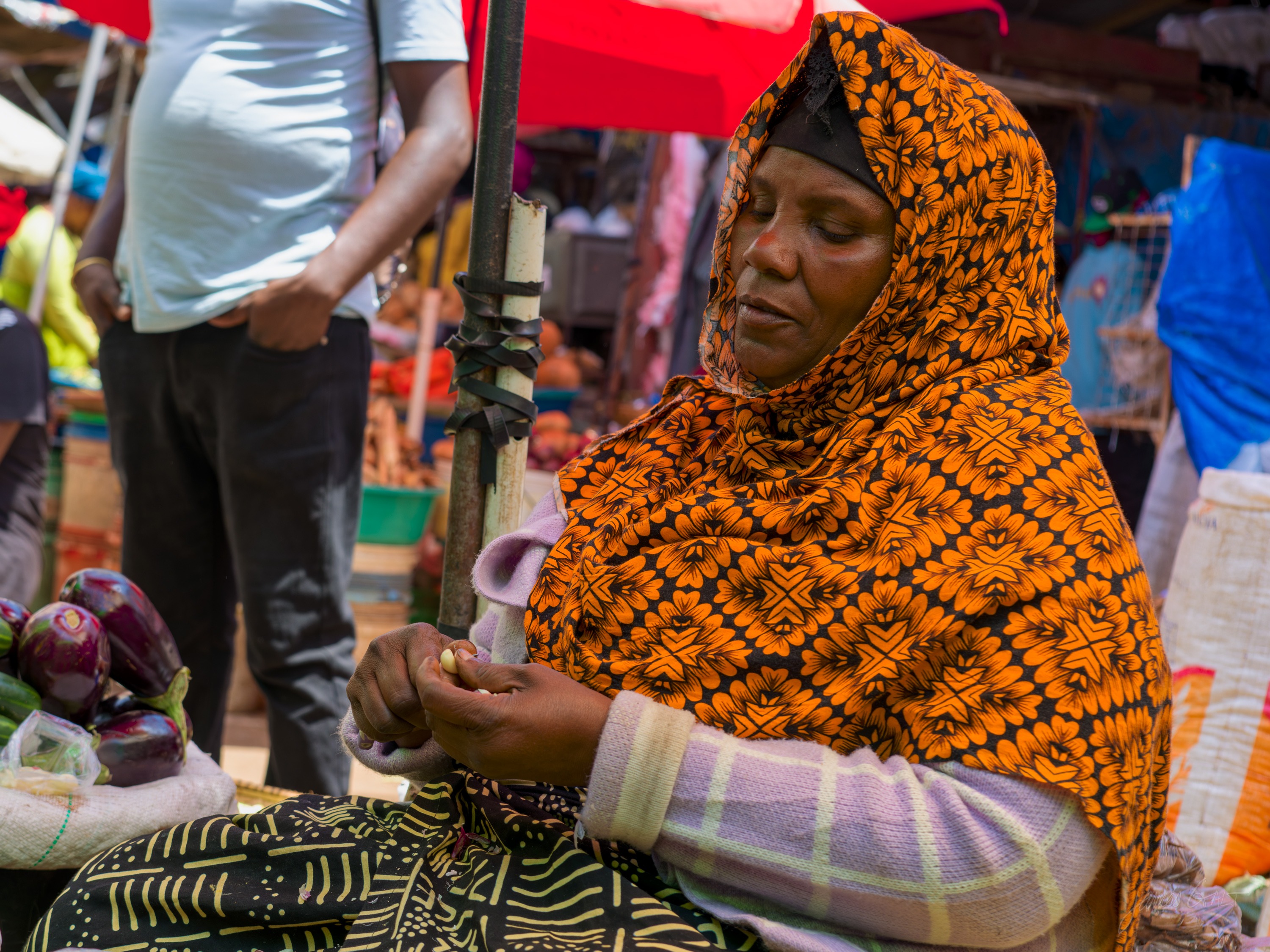 A woman in a patterned orange headscarf and blue top sits at a market stall, looking down at a shallow tray, blurred stalls behind her.
