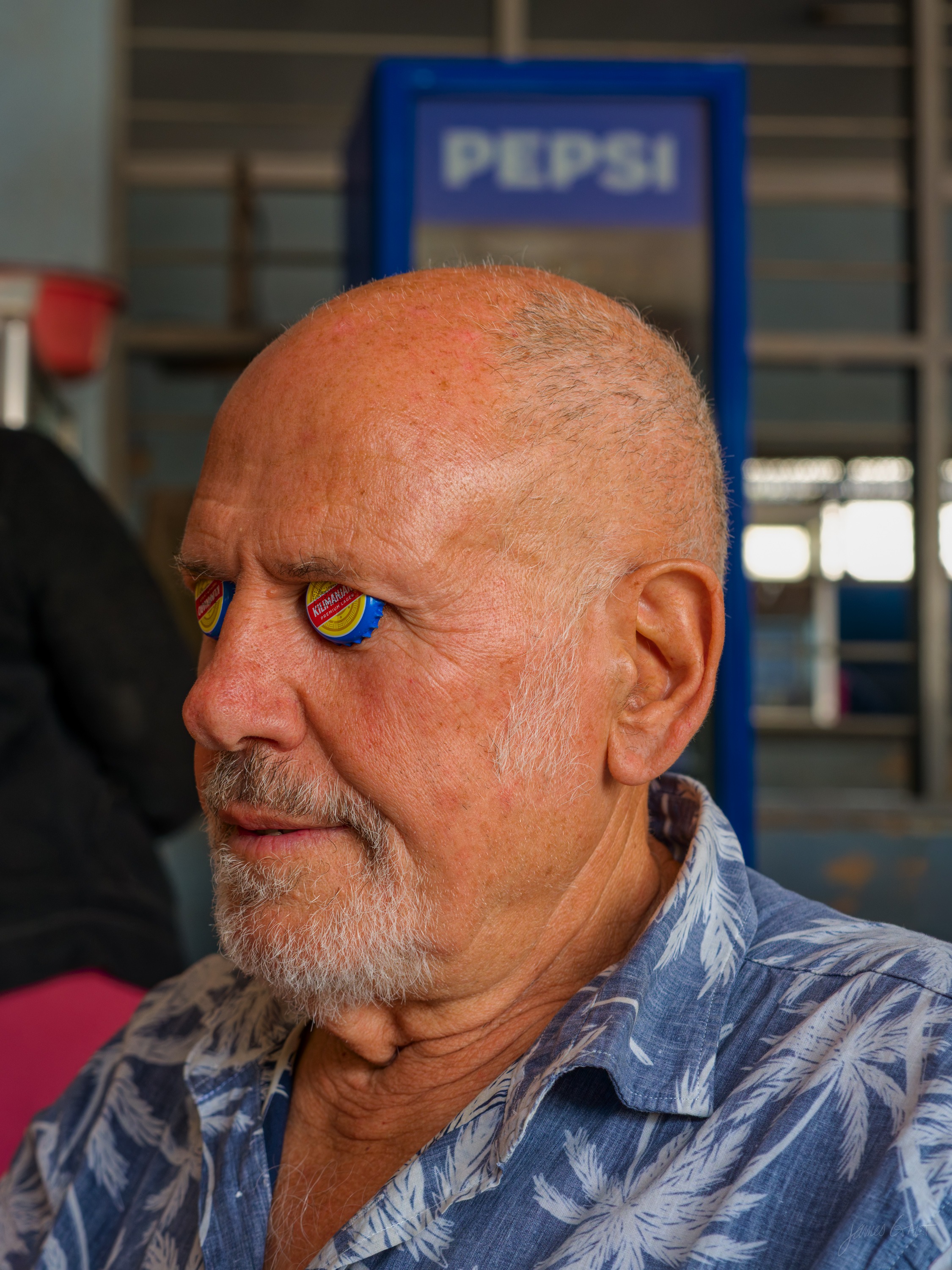 An older man with a grey beard and sunglasses pushed up on his head, in a light patterned shirt, looking to one side beneath a blue Pepsi sign.