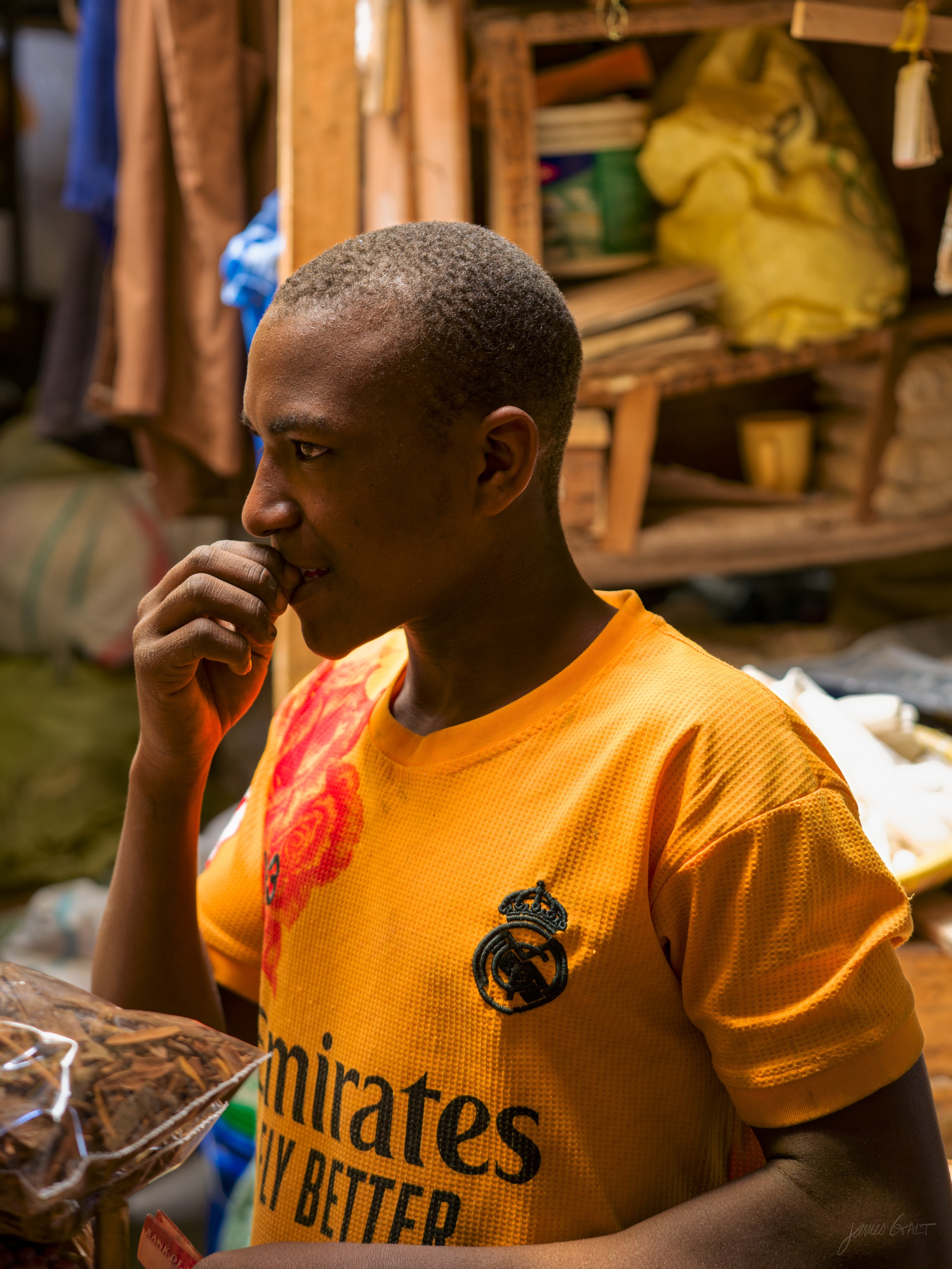 A young man in an orange Emirates football shirt stands among market stalls, one hand near his mouth, looking down in thought.