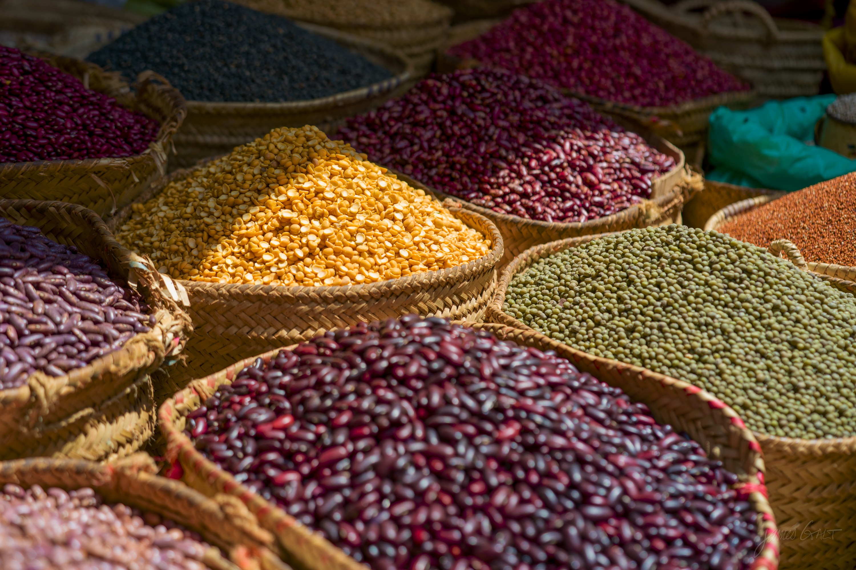Open sacks of dried beans and pulses in many colours arranged in a market stall, seen from above.
