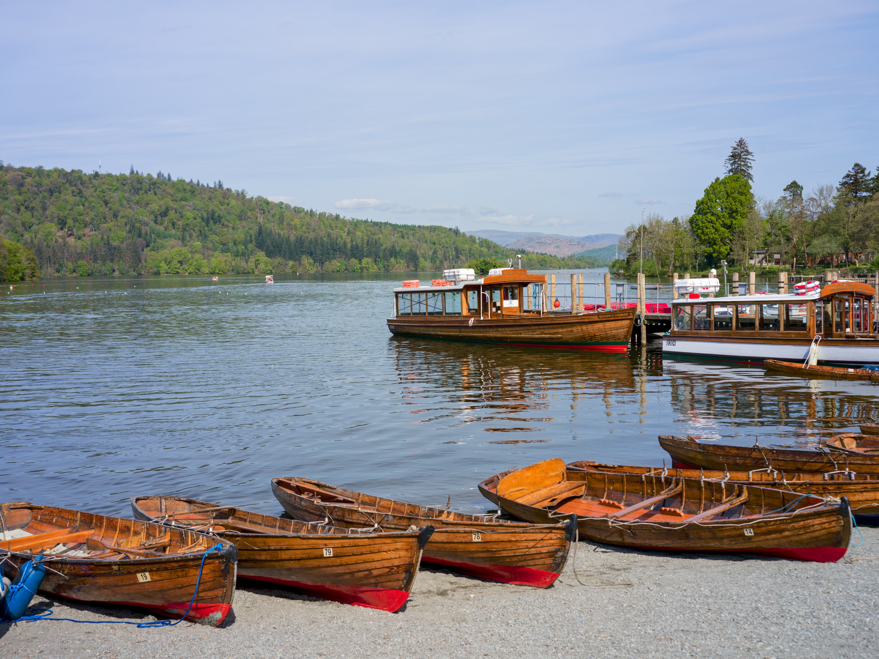 Wooden rowing boats with red keels lined up on a sunlit shingle shore at Bowness, two large wooden launches moored beyond, wooded hills and blue sky across Windermere.