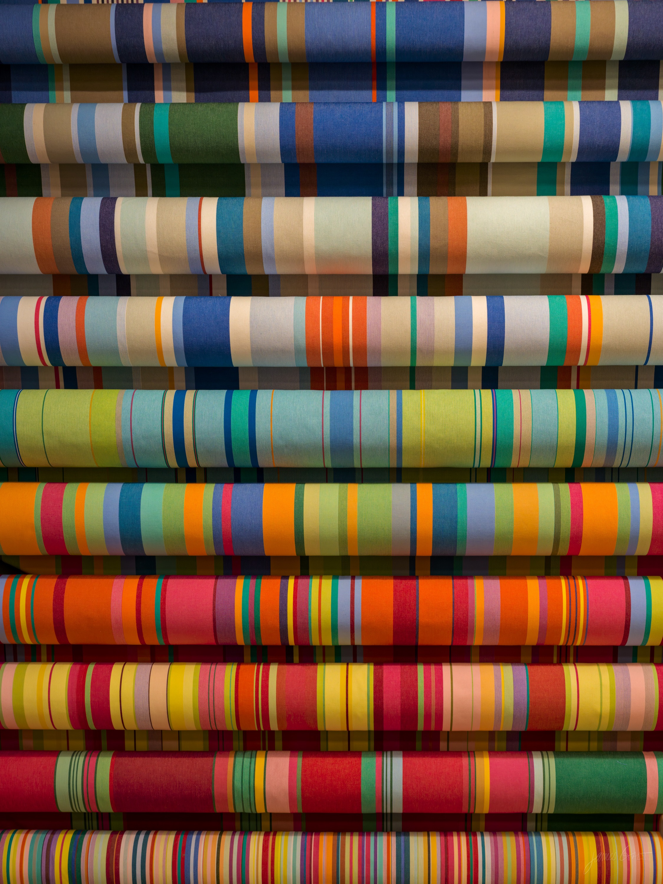 Rolls of woven cotton stacked in rows, each a different multicoloured stripe pattern in bright reds, blues, greens and yellows, seen straight on.