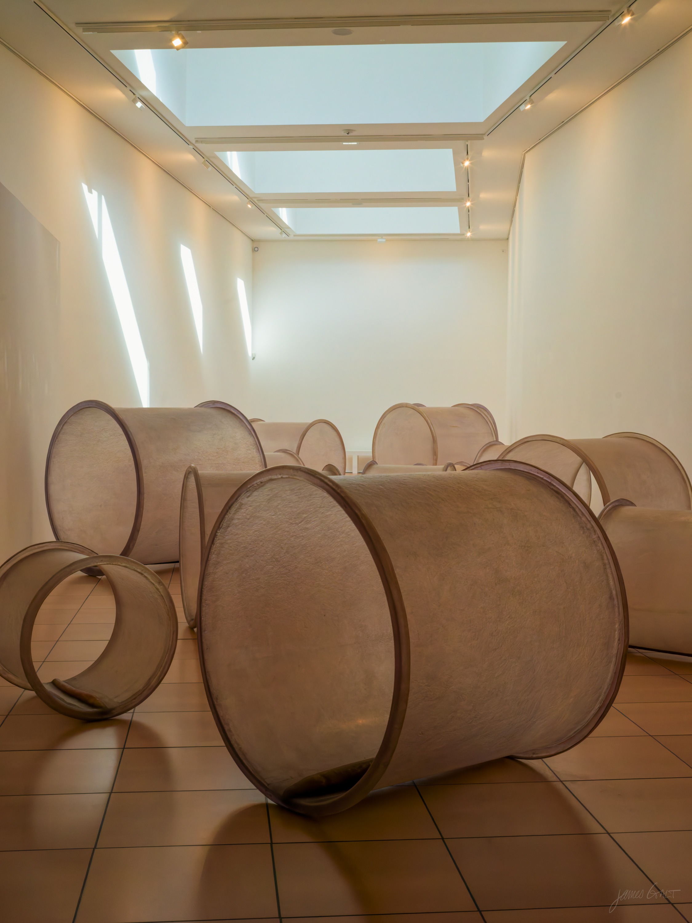 A white gallery room lit by skylights, a group of large pale cylindrical felt sculptures like open drums standing on a tiled floor.
