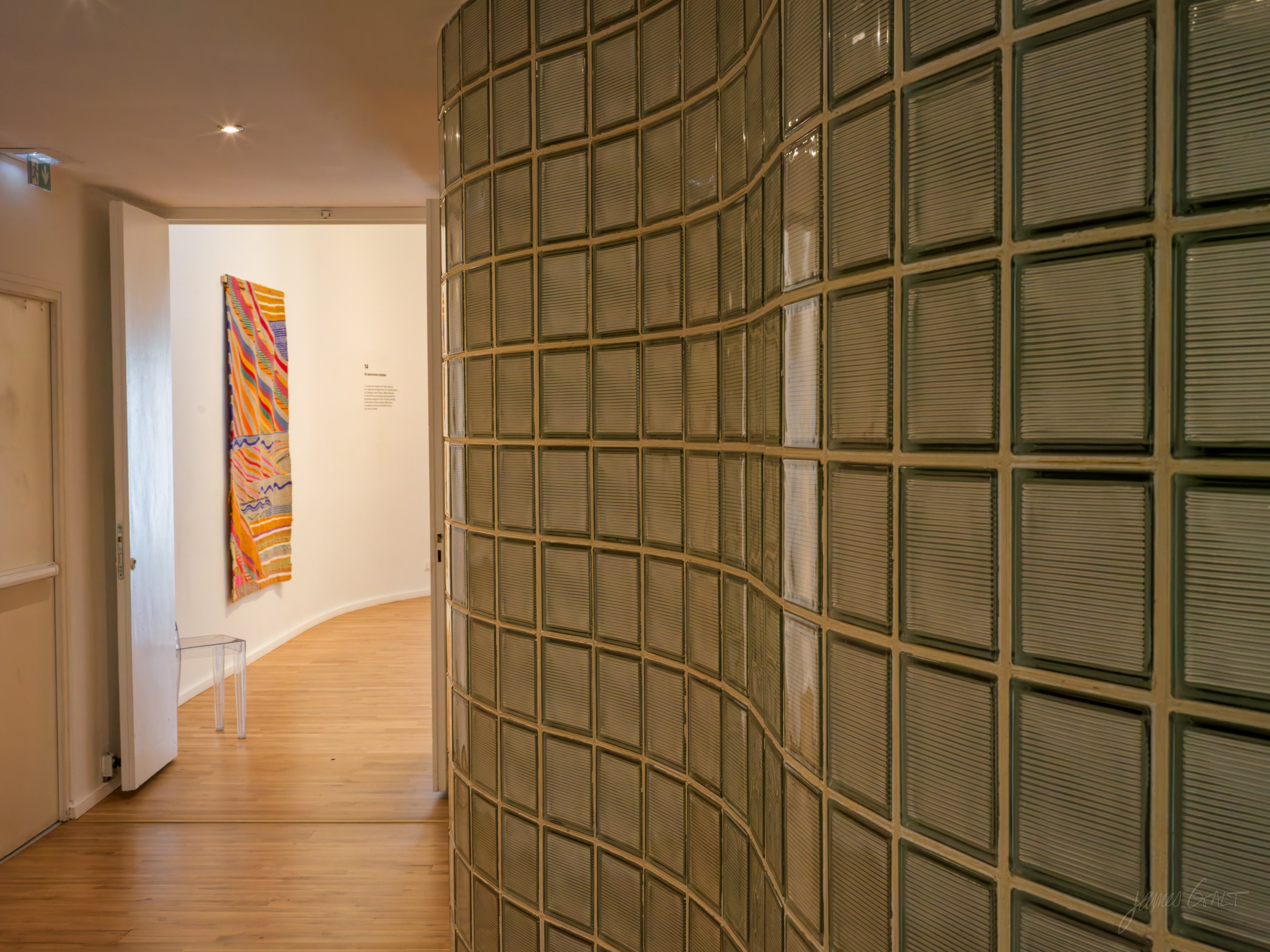 A curved glass-block wall in a gallery, a bright multicoloured woven textile hung on the far white wall beyond it, wooden floor throughout.