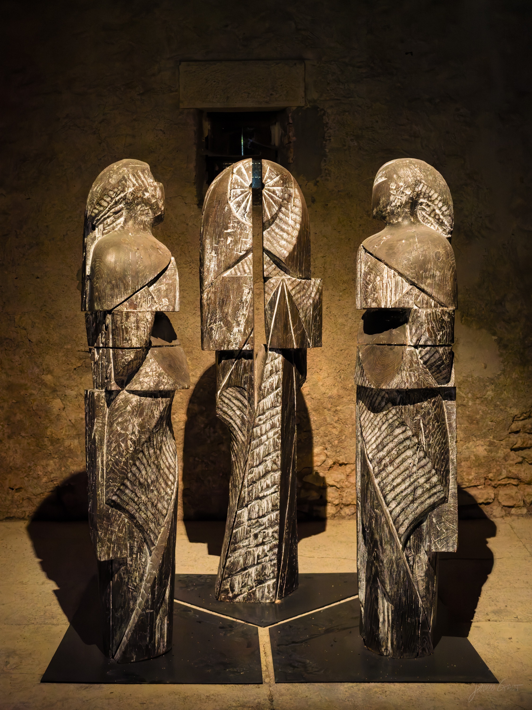 Three tall carved wooden figures on a low plinth in a vaulted stone room, spotlit against the rough wall.