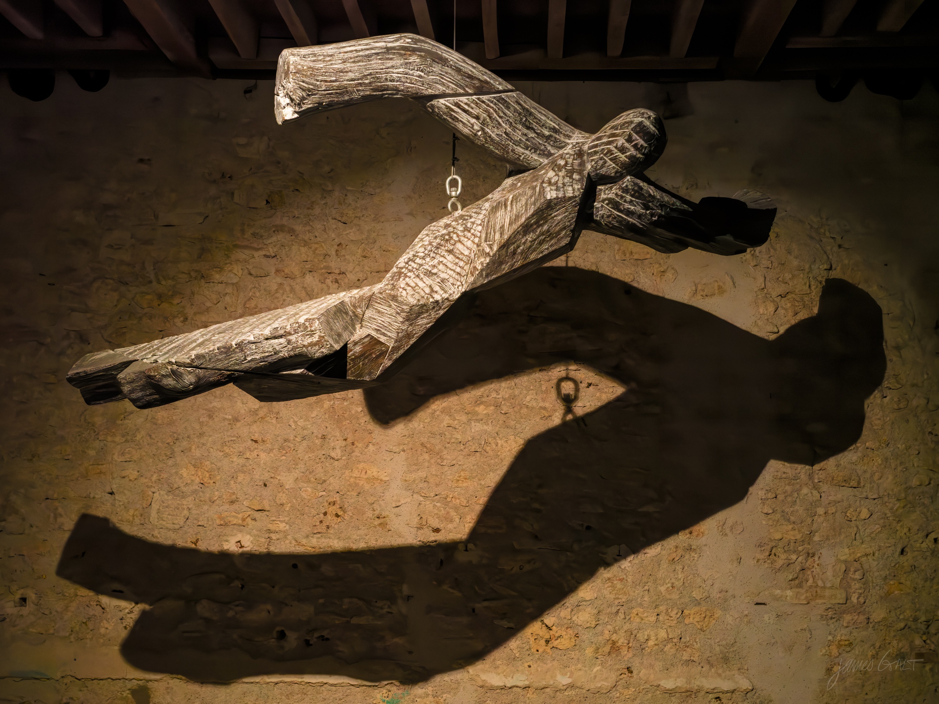 A carved wooden figure with outstretched arms mounted on a stone wall, its exaggerated shadow thrown large beside it.