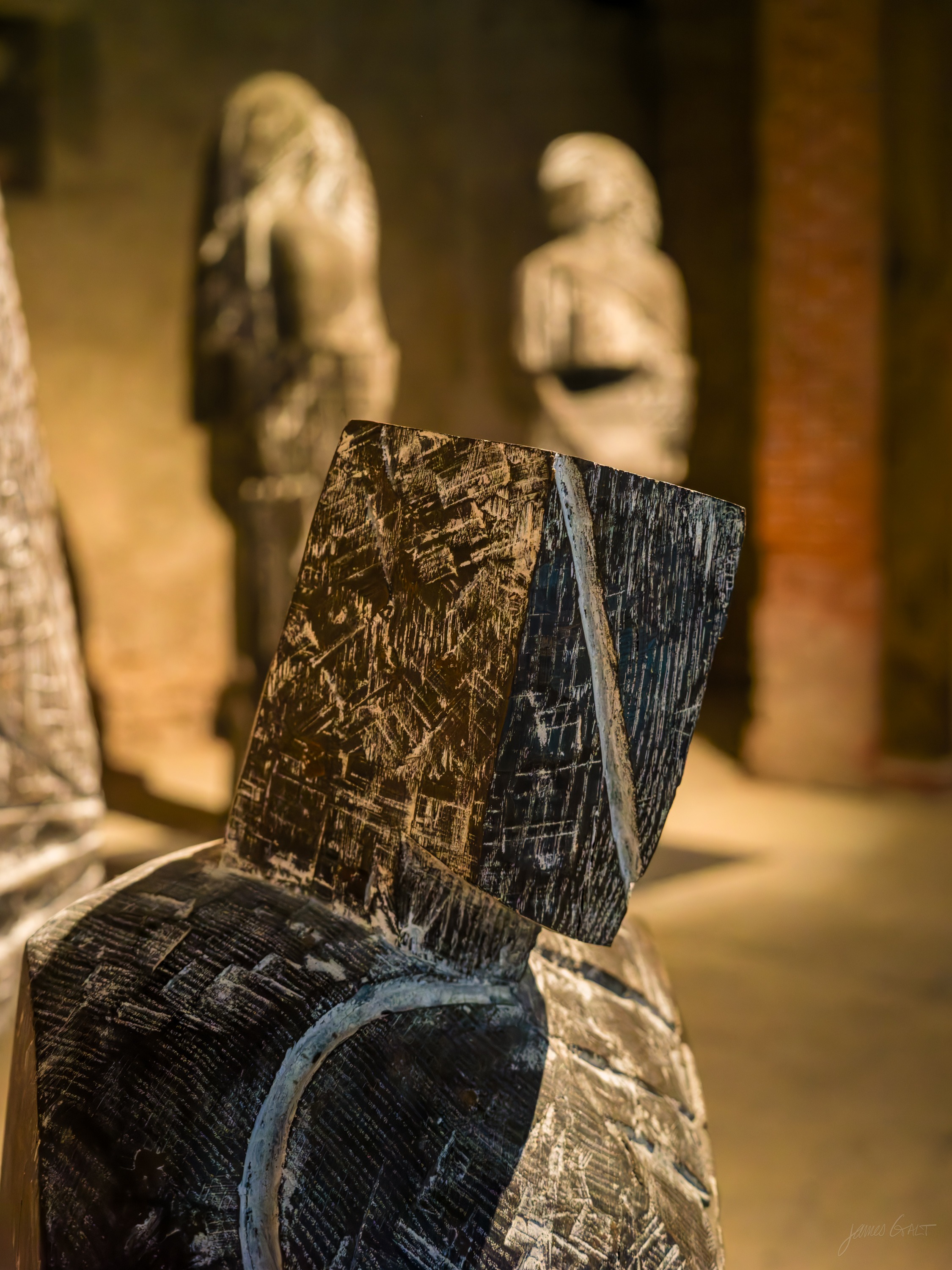 Close view of a split, chisel-marked wooden sculpture in the foreground, two standing carved figures softly blurred behind.