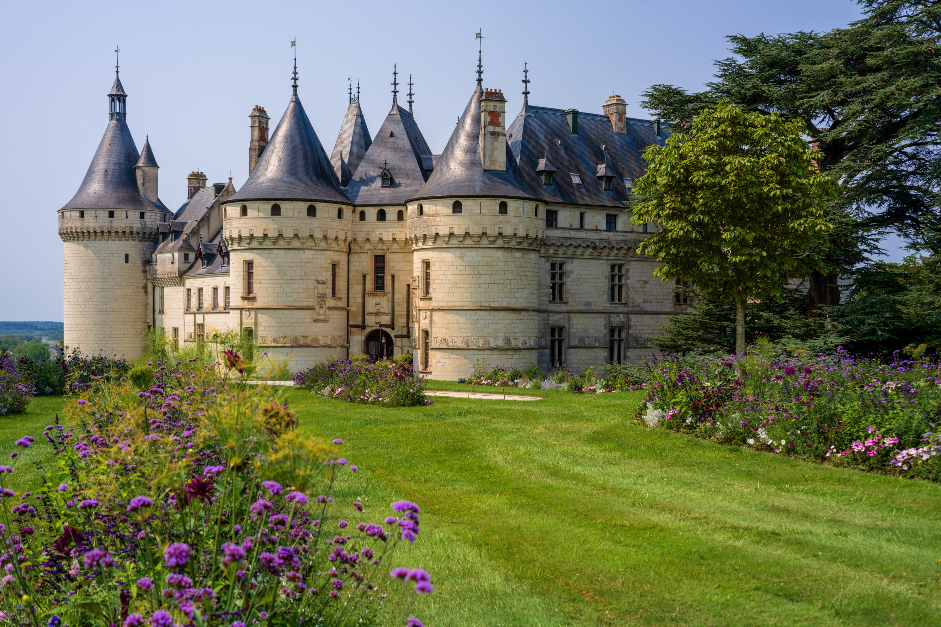 The Chateau de Chaumont-sur-Loire, a pale stone castle with round towers and conical slate roofs, above a lawn edged with purple and mixed flower borders.