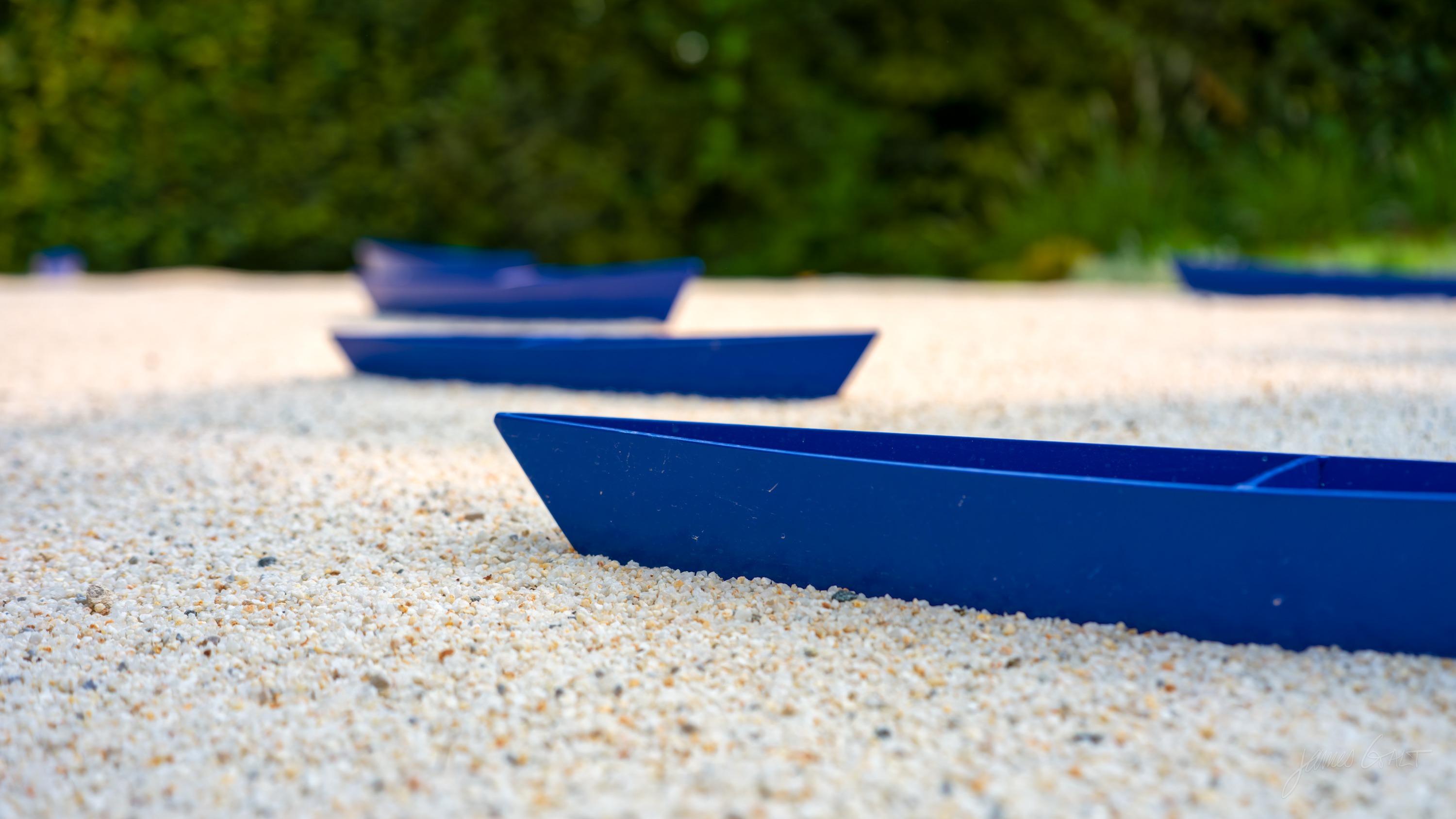 Several small boat-shaped forms in deep blue set on pale raked gravel, the nearest in sharp focus, a green hedge behind.