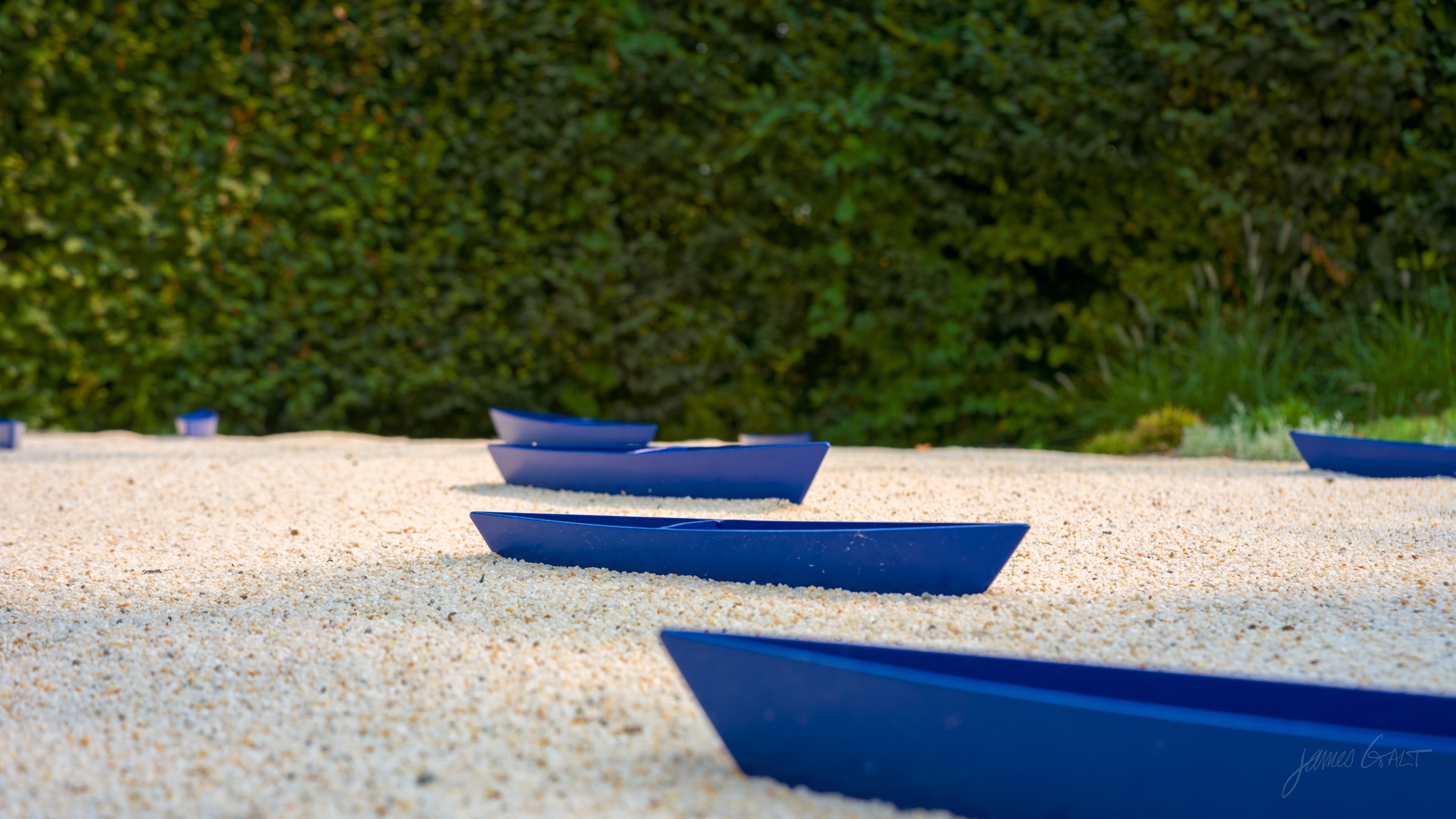 A wider view of the blue boat forms scattered across pale gravel in front of a tall clipped hedge.