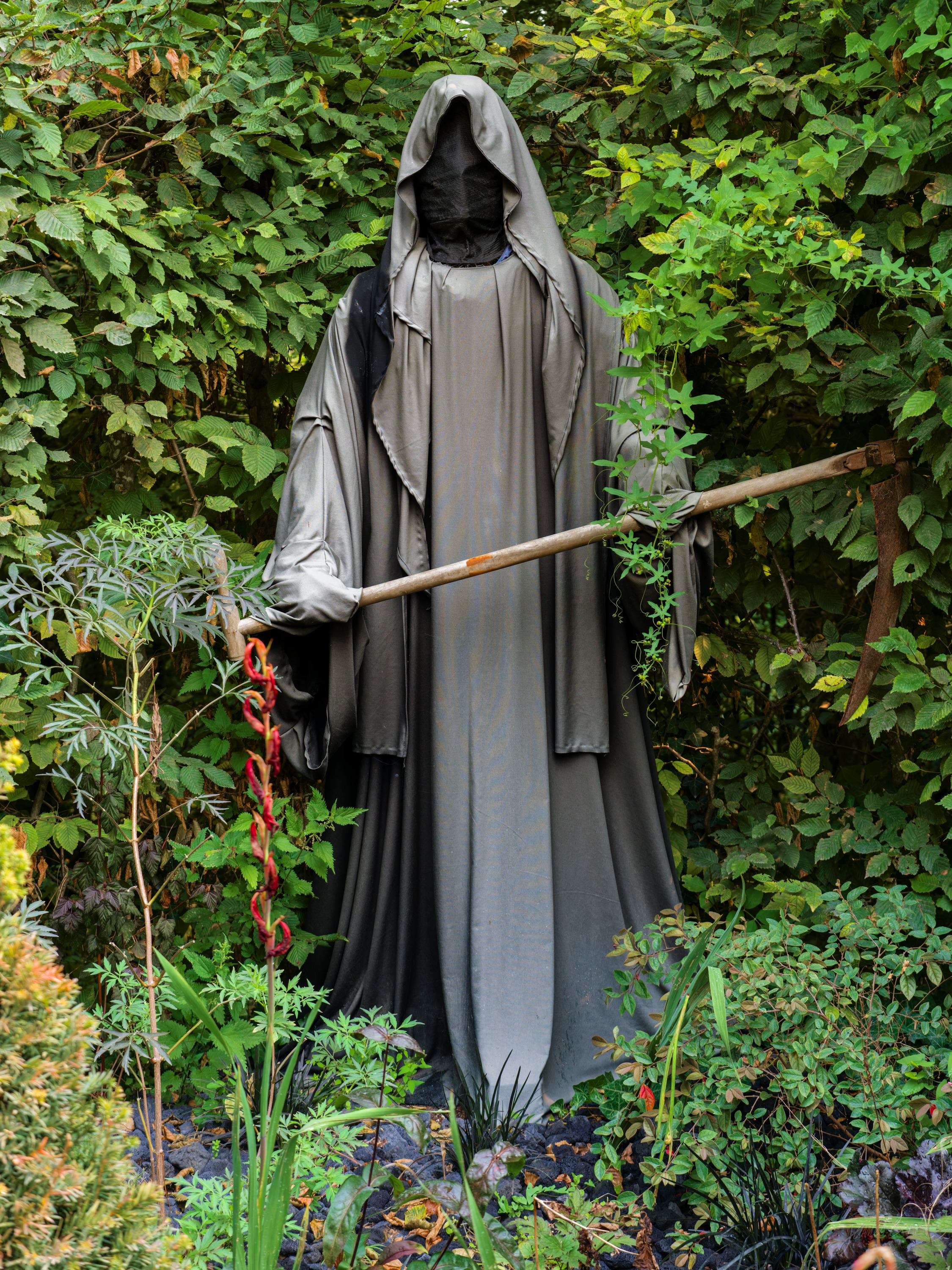 A life-size hooded figure in grey robes standing among dense green garden planting, face in shadow, holding a staff.
