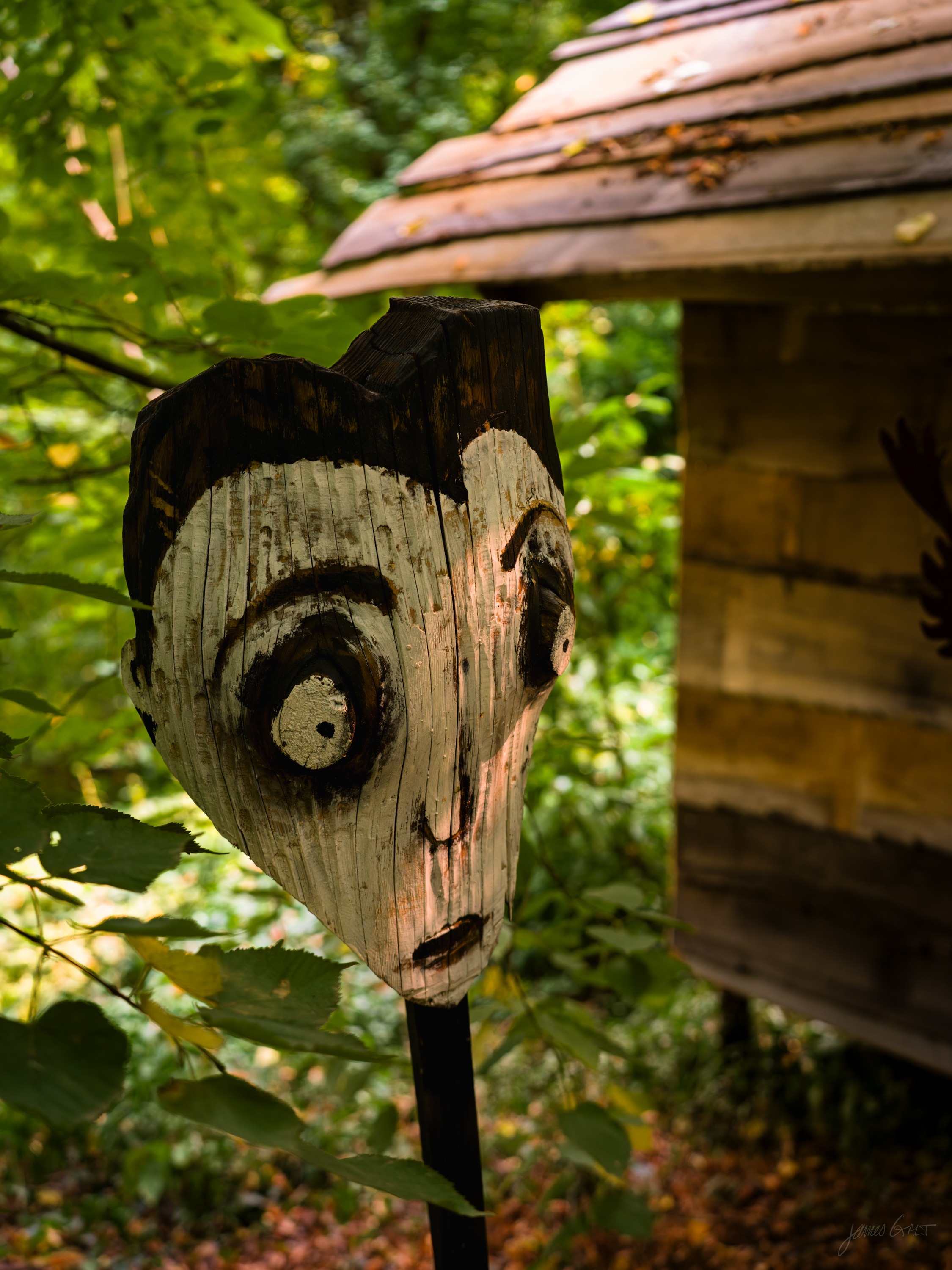 A carved wooden mask with wide painted eyes mounted on a post, a small timber hut behind it in green woodland.