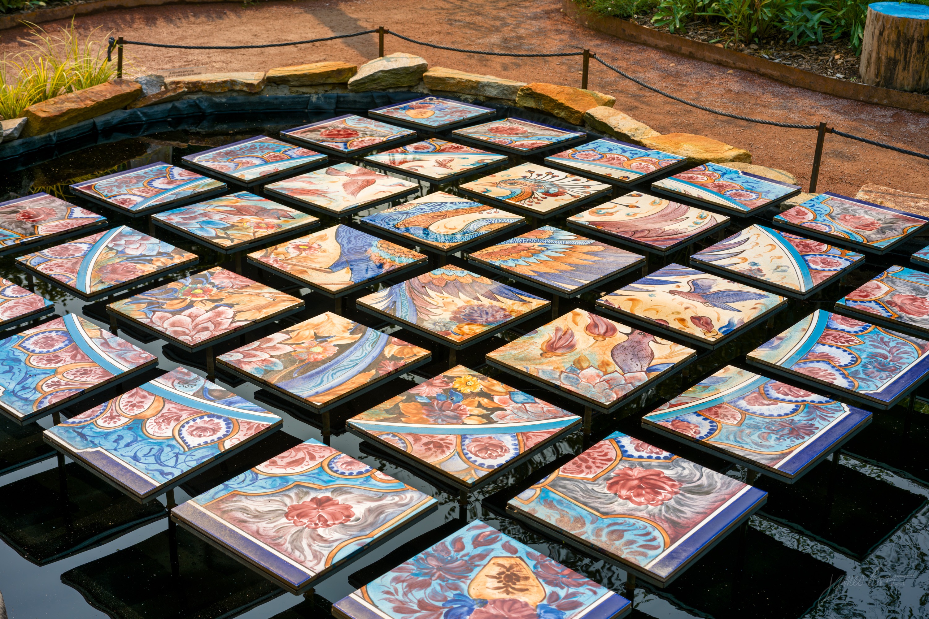 A grid of square painted ceramic tiles floating on a dark rectangular pool, each tile patterned in blues, reds and ochres.