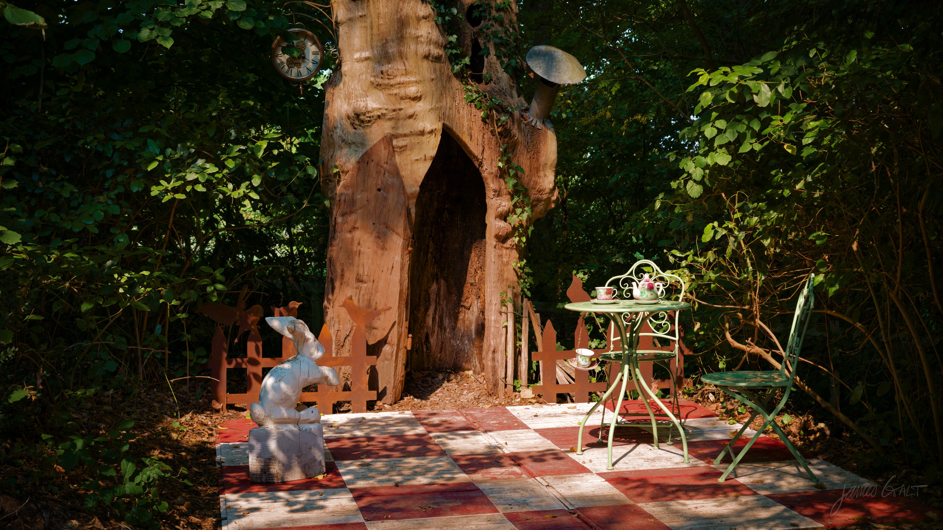 The hollow trunk of a large tree opening onto a red-and-cream checkerboard floor, with a pale deer figure and an ornate metal cafe table and chairs.