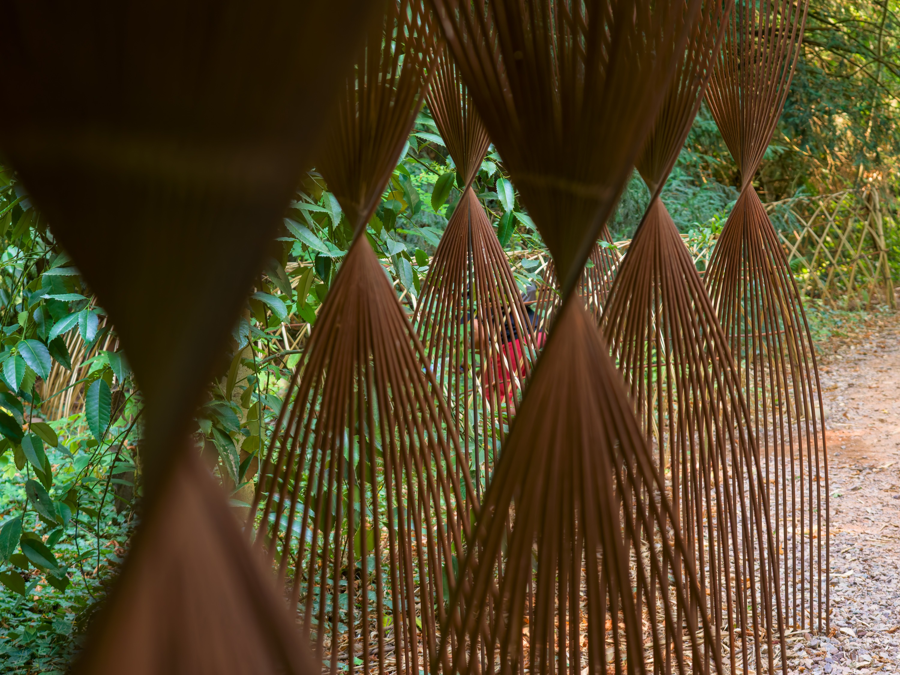 A hanging screen of woven cone shapes in rust-brown, strung in rows across a woodland path.