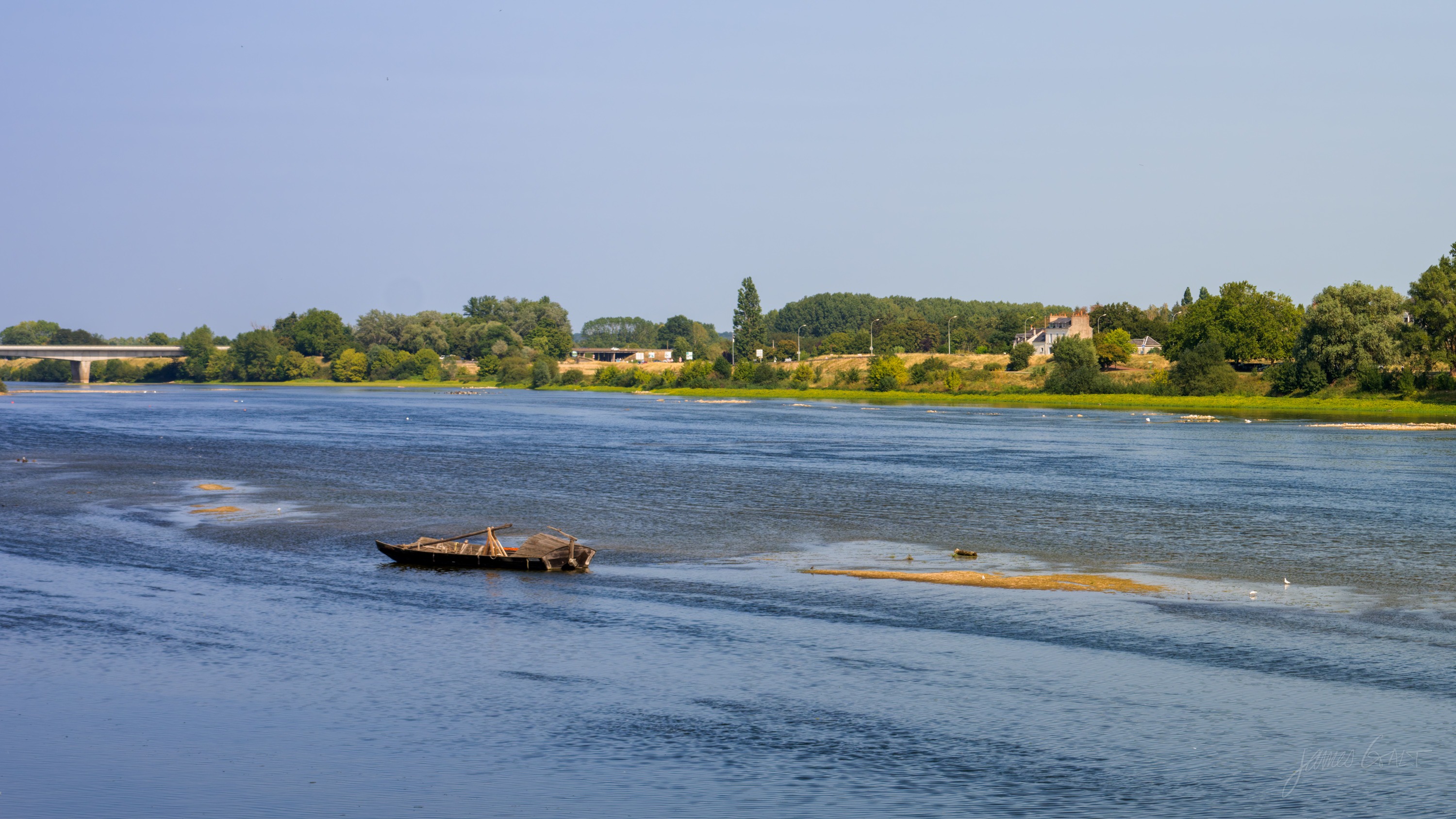A wide river under a pale sky, exposed sandbars breaking the surface, a single traditional flat-bottomed wooden boat moored mid-channel, a low bridge and wooded bank in the distance.