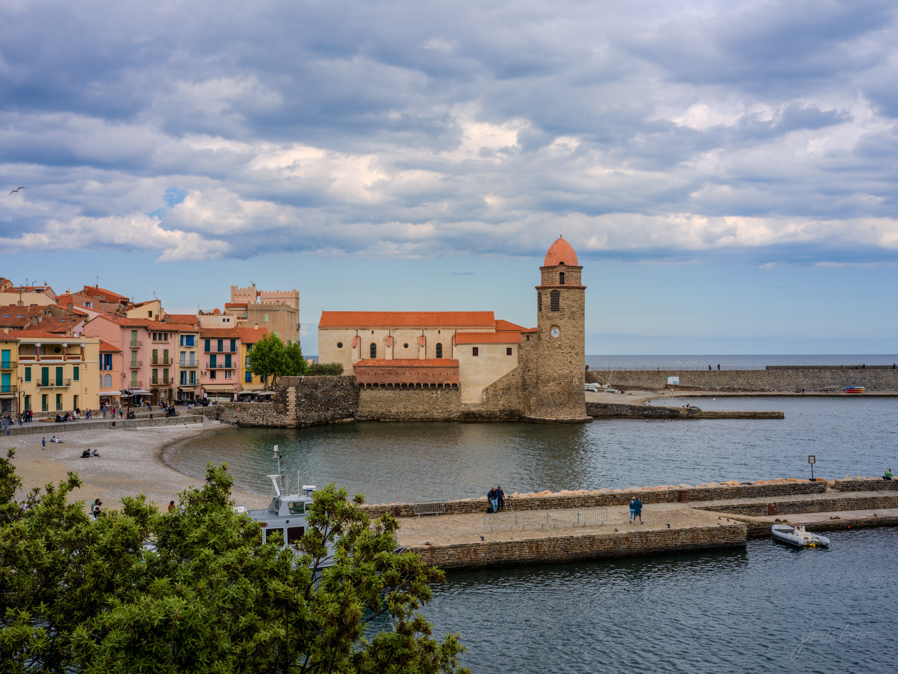 A terracotta-roofed town wrapped around a bay, a wooded hill behind topped by a small star fort, vineyards on the slopes, blue sky with cloud.