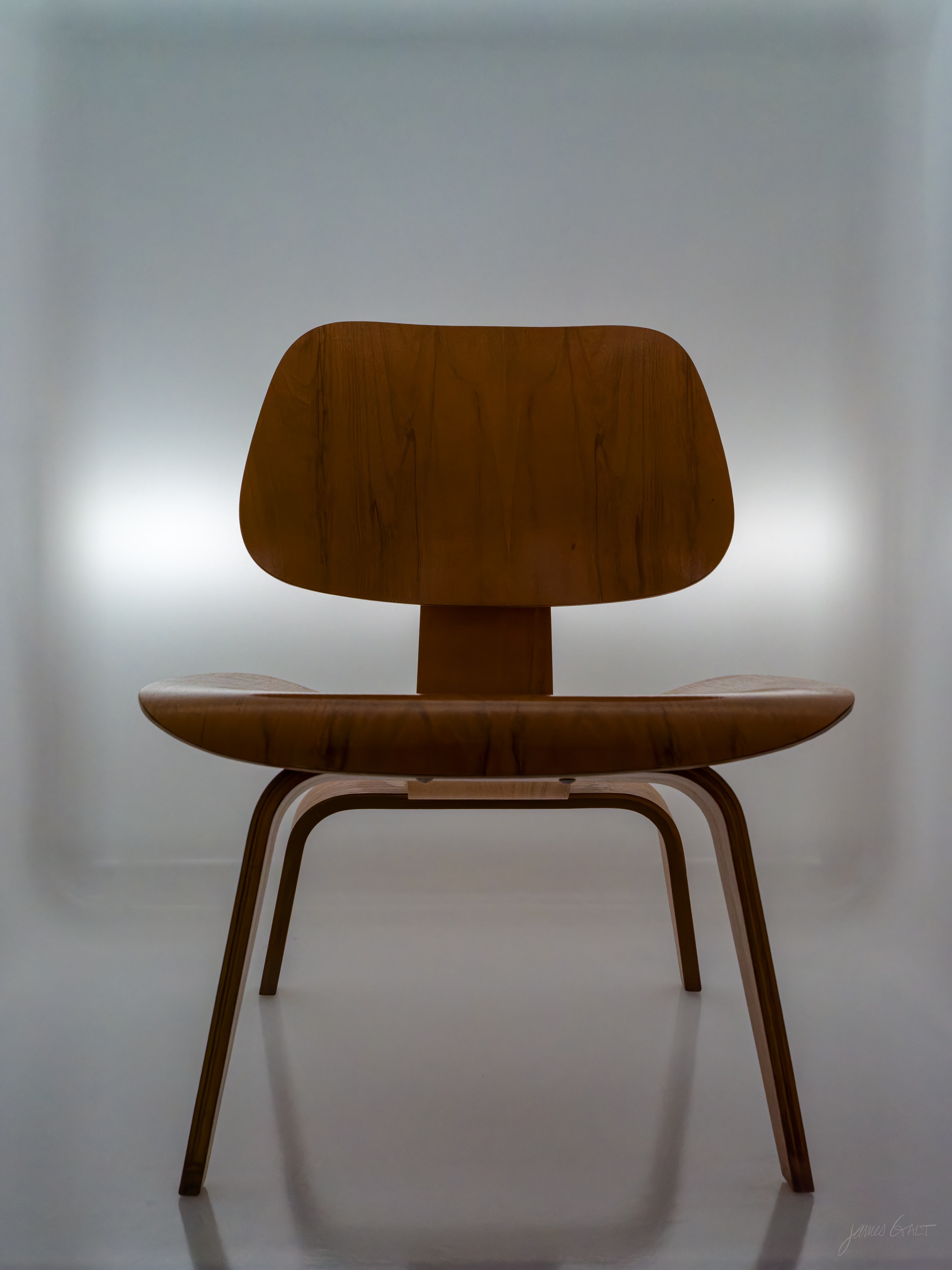A molded plywood chair with a curved back and seat in warm honey-toned wood, photographed straight on and centered against a glowing white backlit panel, its legs reflected on the pale floor.