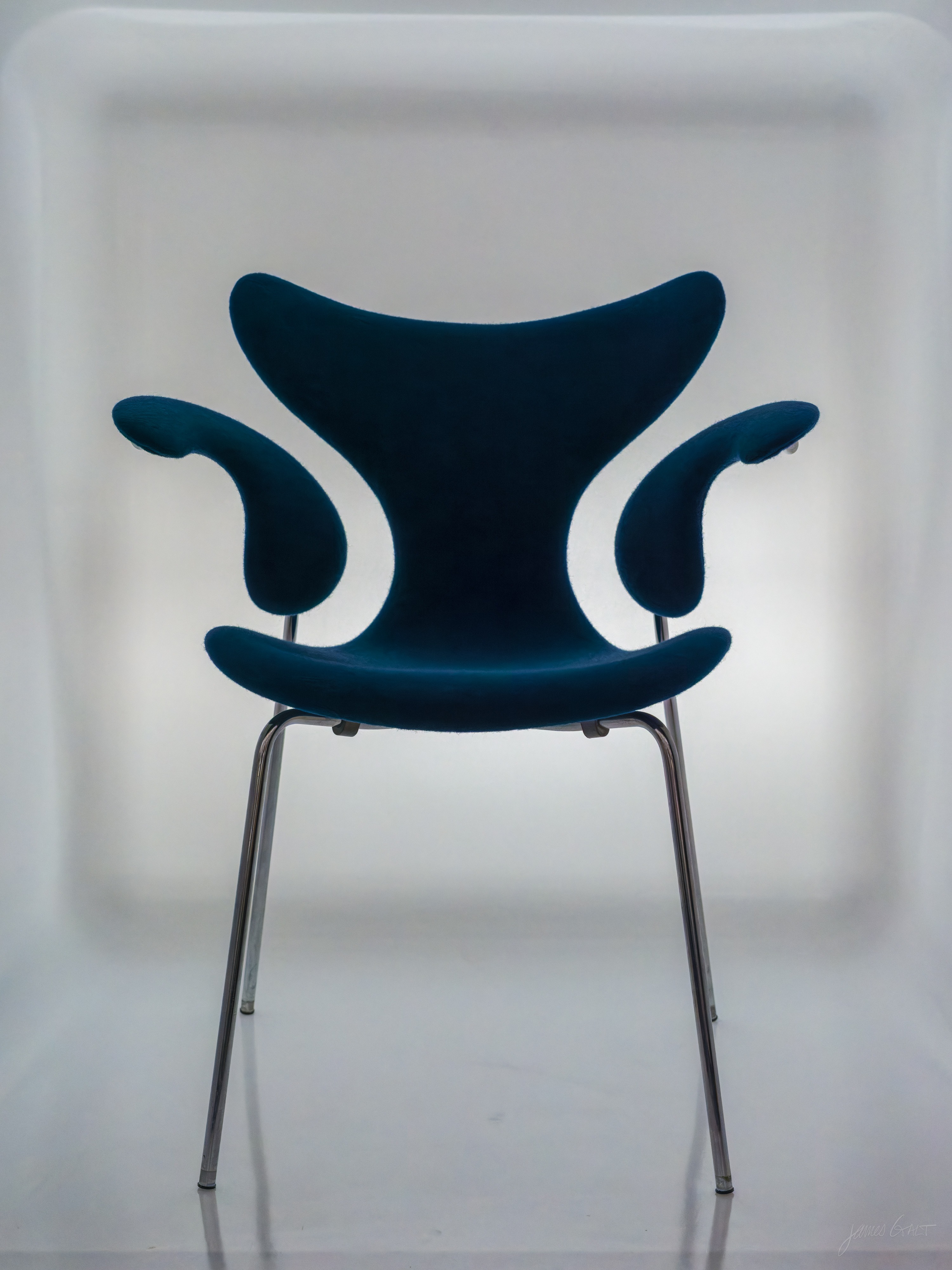 A blue upholstered armchair with a sculpted back and distinctive curling, horn-like armrests on slender chrome legs, photographed straight on against a glowing white backlit panel.