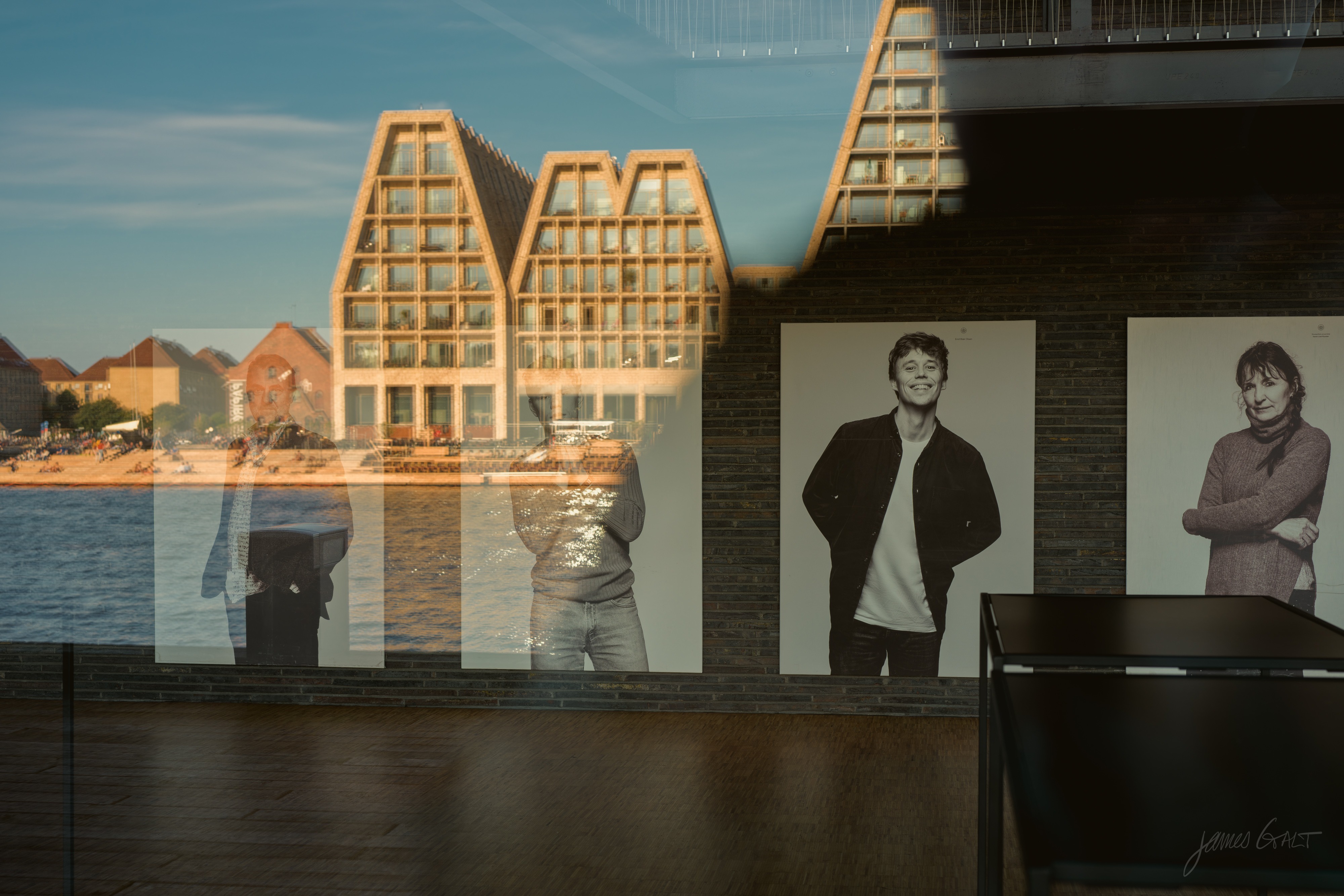 Layered reflections in a window: a row of angular-roofed waterfront buildings and the harbor mirrored across the glass, behind which two large black-and-white portrait photographs, a young man and a woman, hang on a brick wall above a wooden floor.