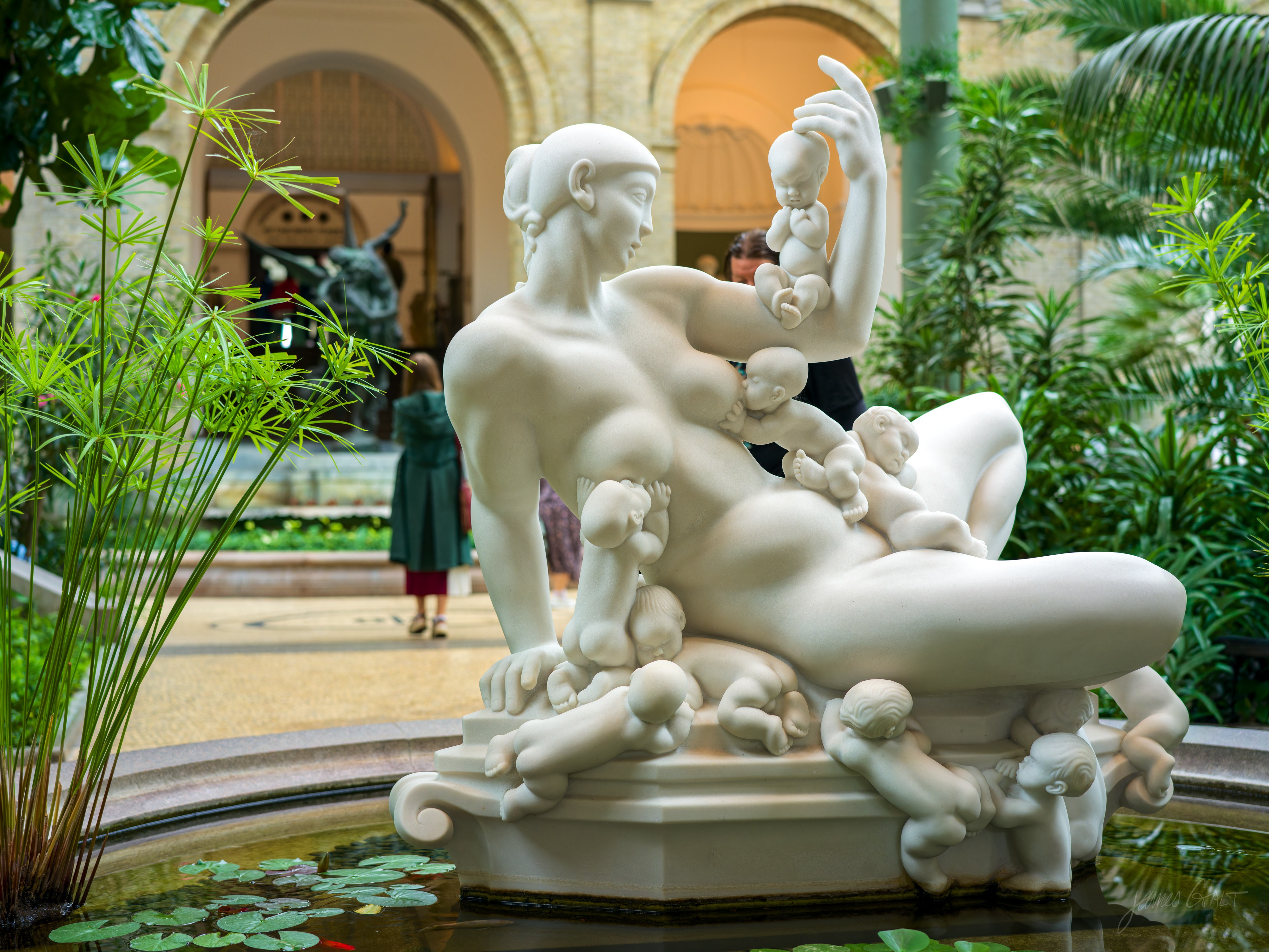 A white marble sculpture of a reclining woman with many small babies climbing over her body, on a fountain base ringed by lily pads, surrounded by palms and greenery, with blurred visitors in the arched gallery behind.