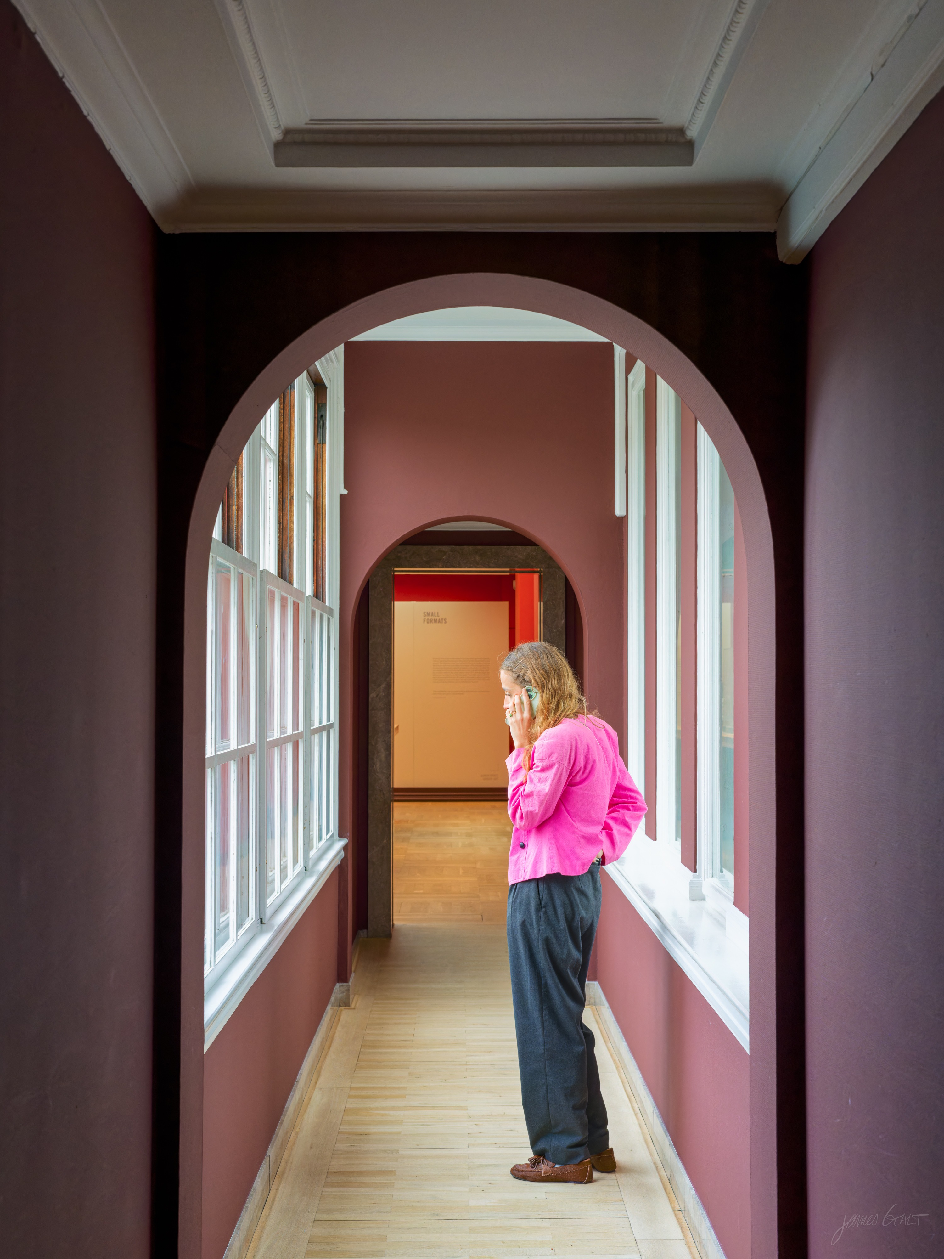 A museum corridor with deep rose and maroon walls and rounded arches receding into the distance; a person in a bright pink shirt and dark trousers stands in profile by a window, on the phone, with a doorway to an orange-lit room at the far end.