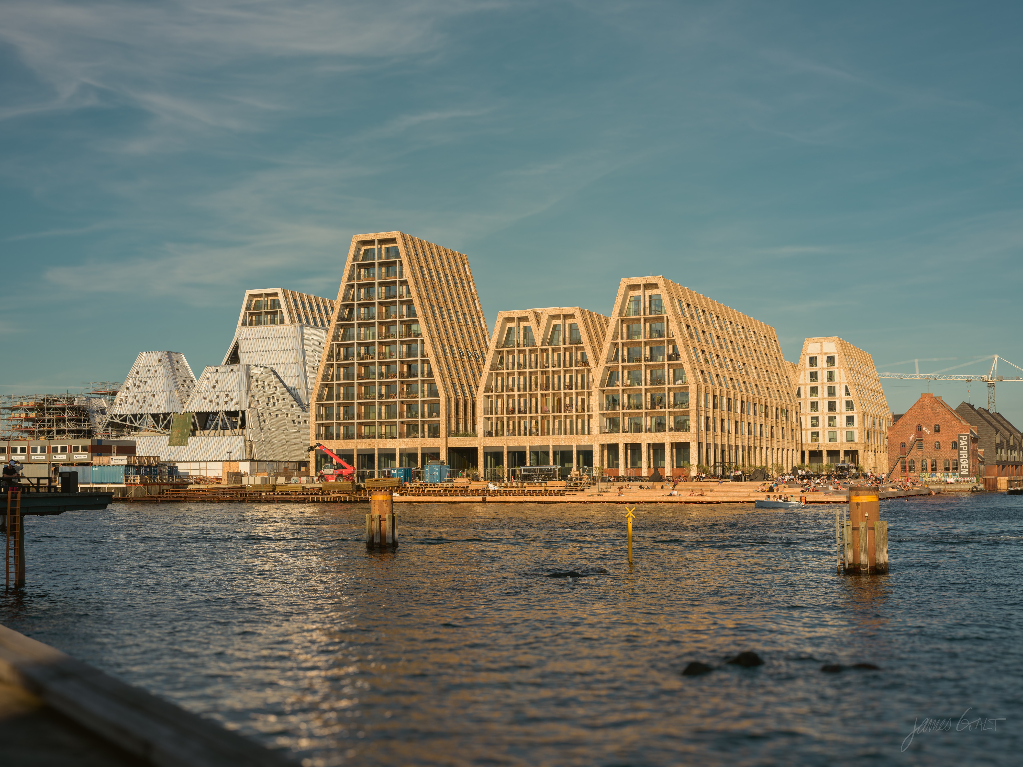 A row of modern apartment buildings with steep, angular gabled roofs on the Christianshavn waterfront, lit gold by low evening sun and seen across calm harbor water, with old brick warehouses to the right and a white faceted building to the left.