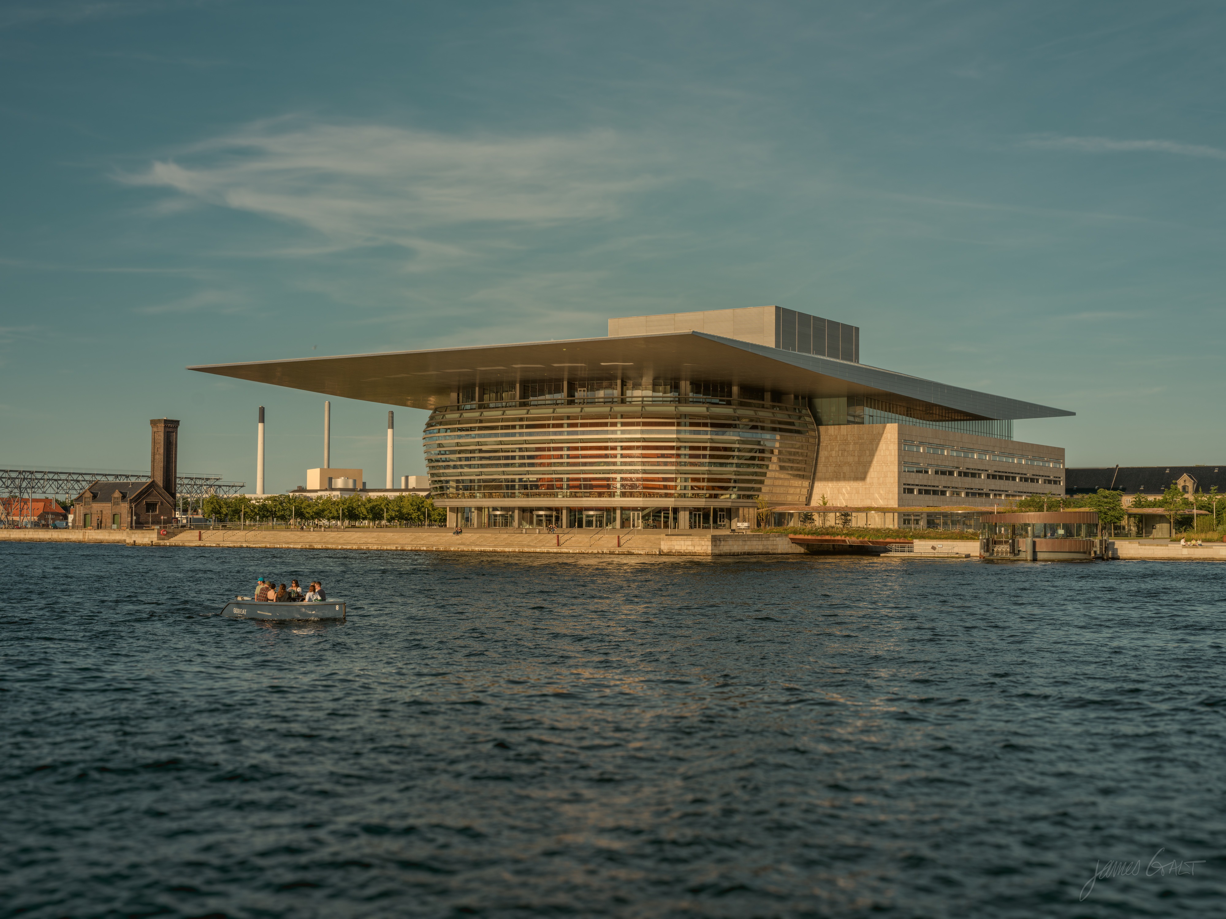 The Copenhagen Opera House across the harbor, its broad flat roof cantilevered over a curved glass front, with a small open boat carrying a few people in the foreground and industrial chimneys on the skyline behind.
