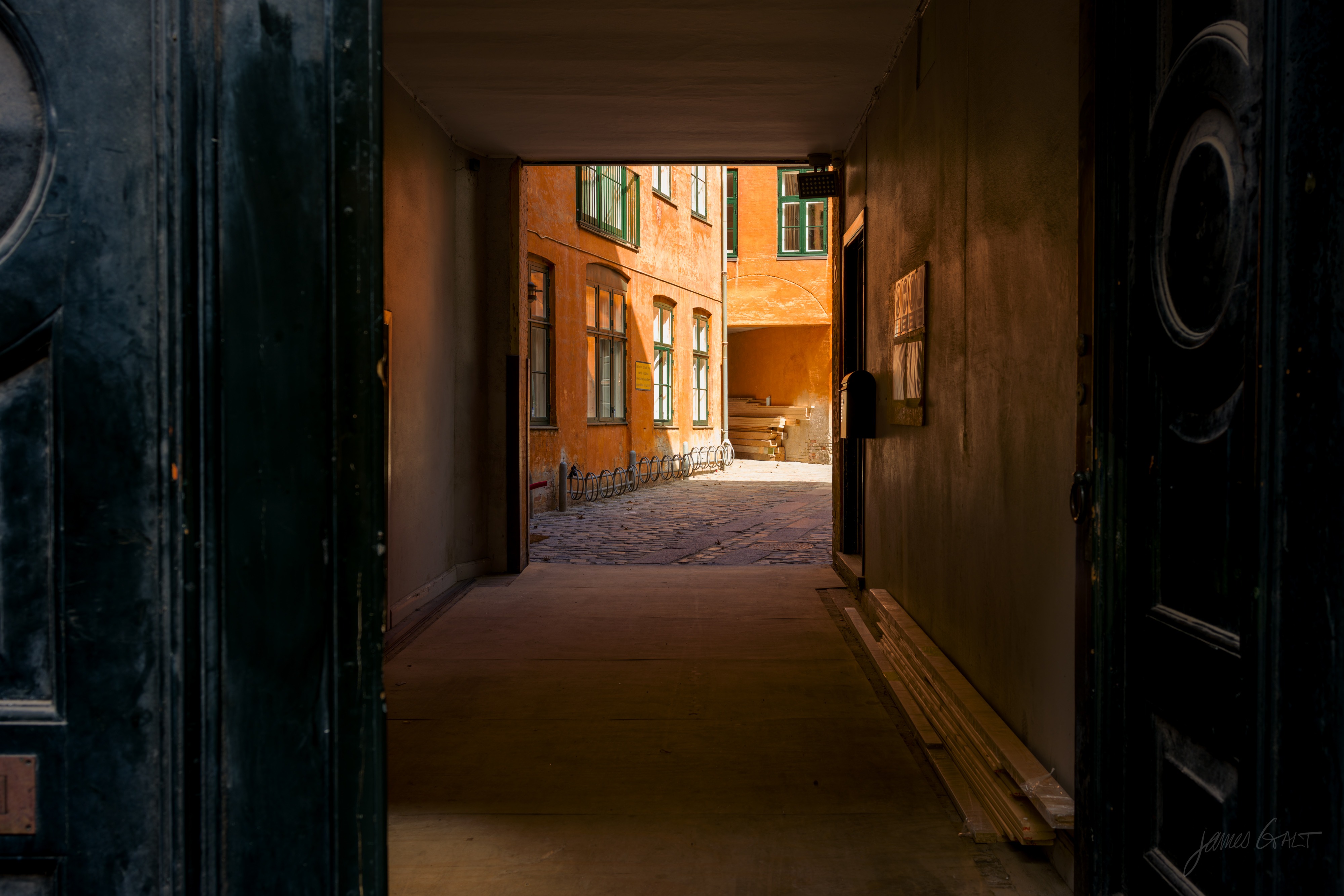 View through a dark stone gateway out to a sunlit courtyard: an ochre building with green-framed windows, a curved bicycle rack, and cobblestones beyond.
