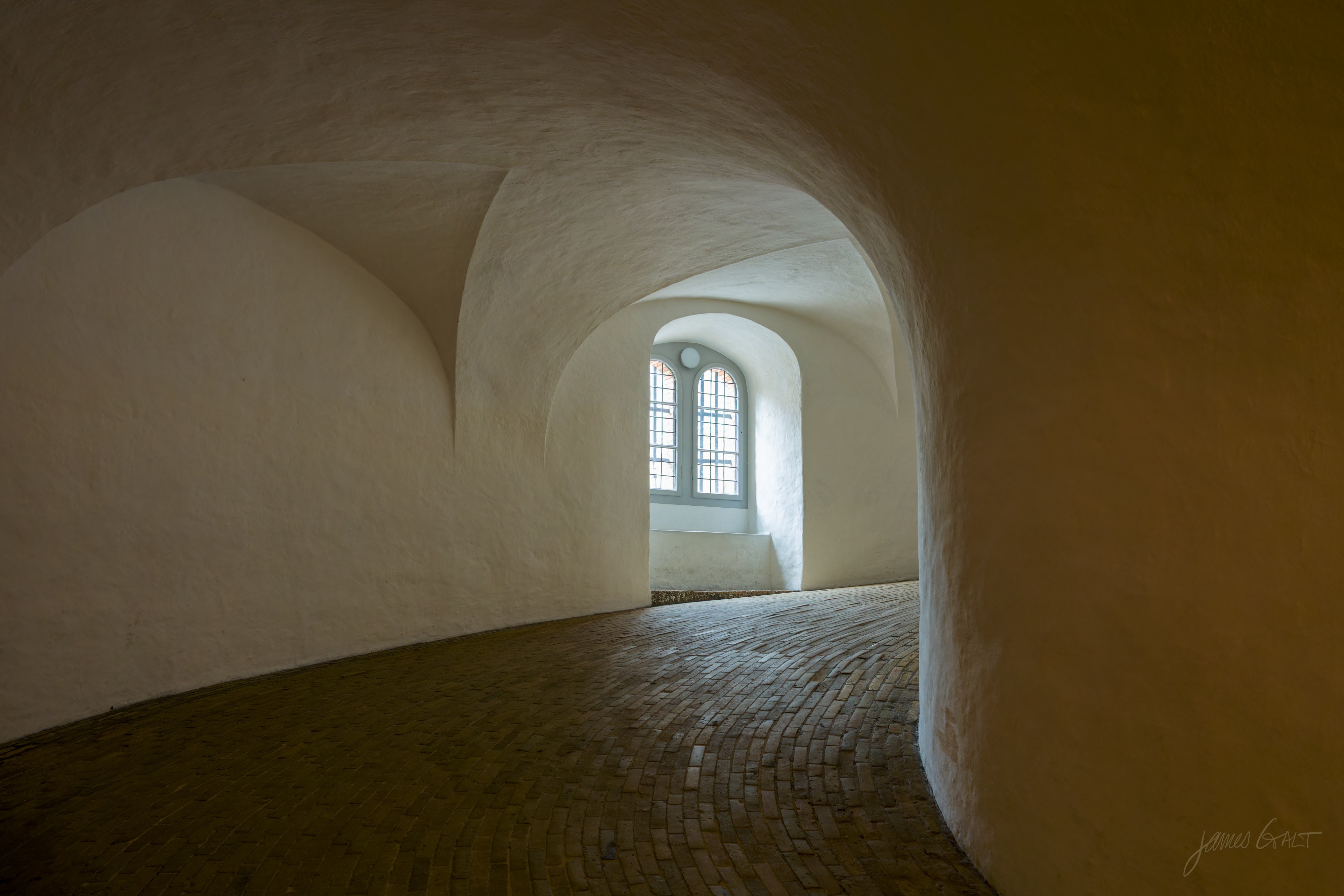 Interior of the Round Tower (Rundetarn) in Copenhagen: a spiral masonry ramp with whitewashed vaulted walls and a worn brick floor curving away toward a tall arched window letting in pale light.