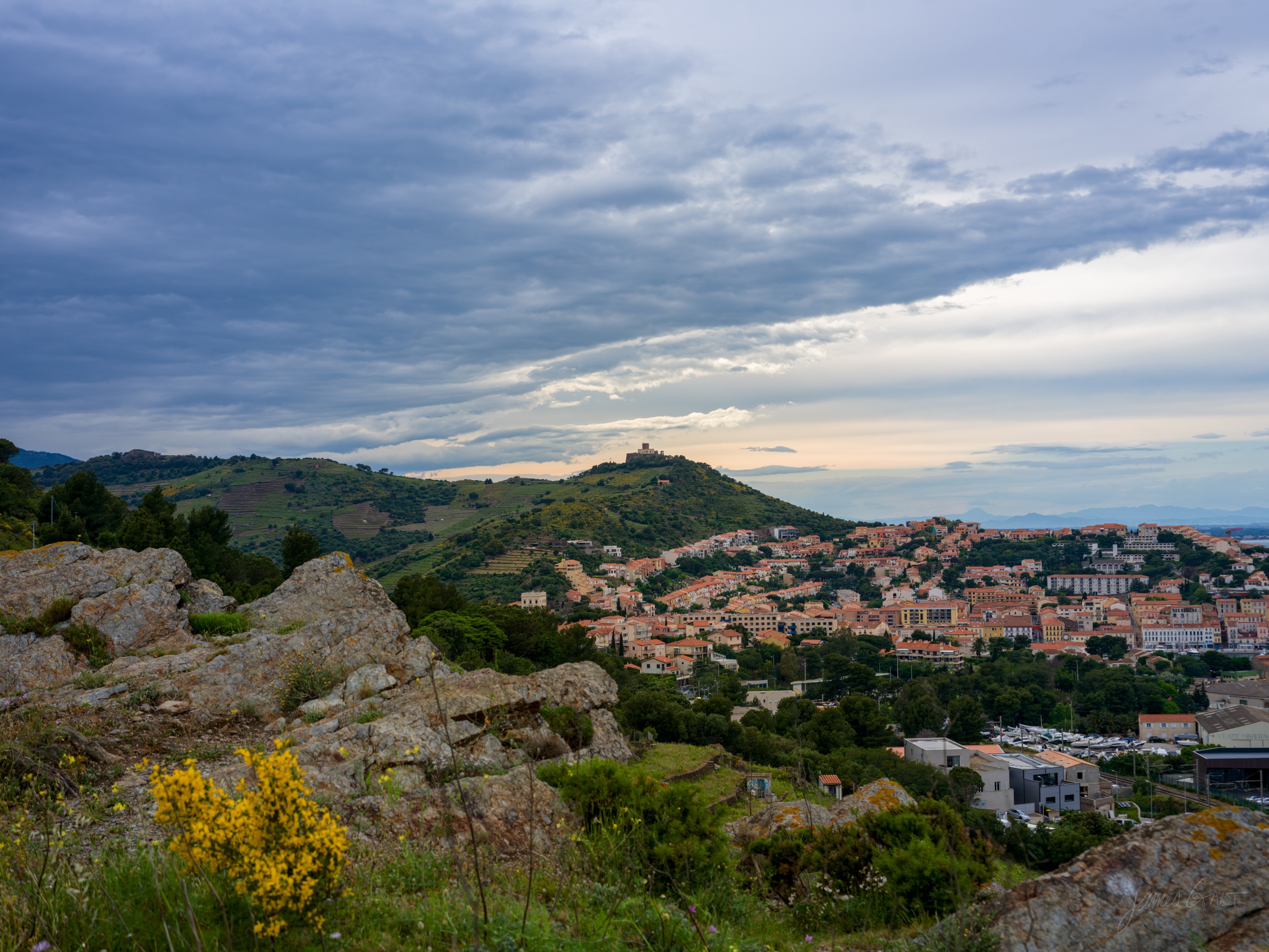 A hillside town of terracotta roofs seen from rocky ground across a valley, a small fort on the ridge behind, warm evening light breaking under grey cloud.