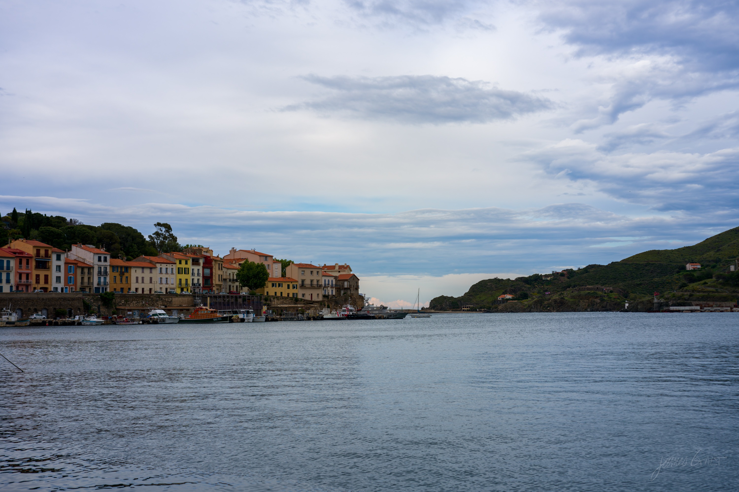 A harbour of moored boats in front of a terrace of pastel buildings on the far quay, wooded headlands closing the bay under a broad soft sky.