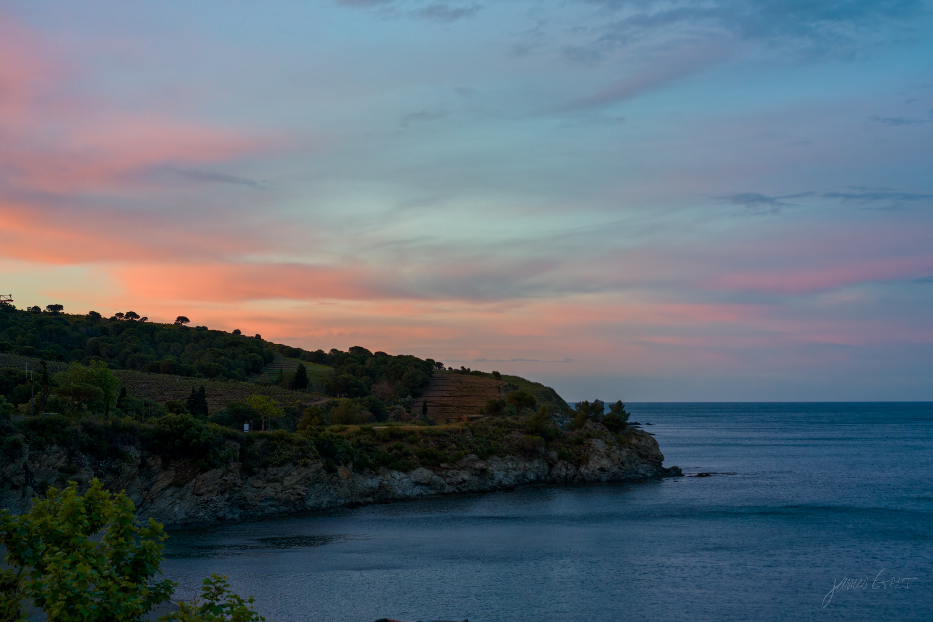 A pink and orange sunset over a low headland and a calm cove, the sun's warmth breaking beneath a band of cloud, the sea gone dark below.
