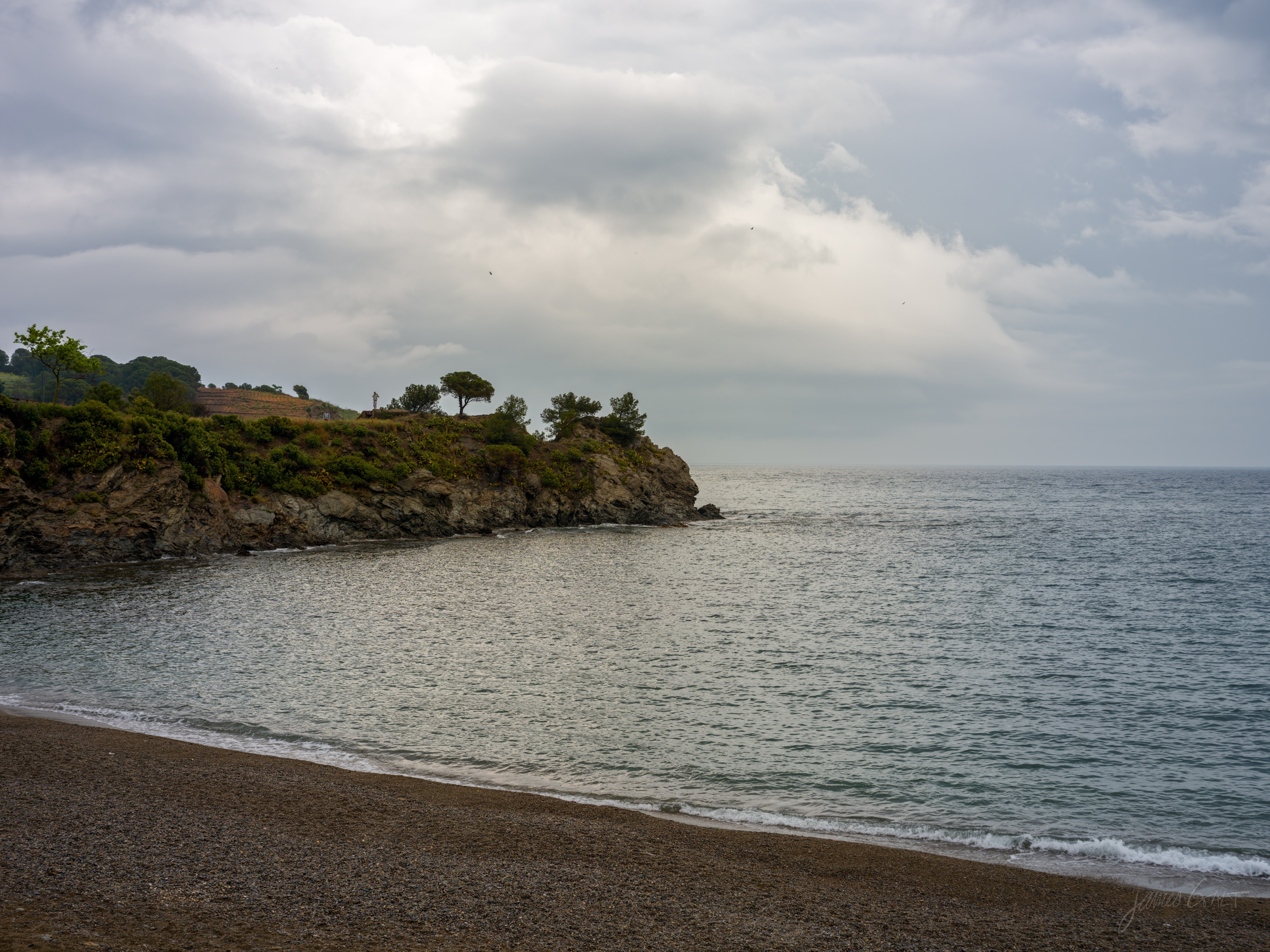 A pebble cove with a low rocky headland topped by umbrella pines, a flat grey sea and heavy cloud beyond, calm water lapping the shingle.