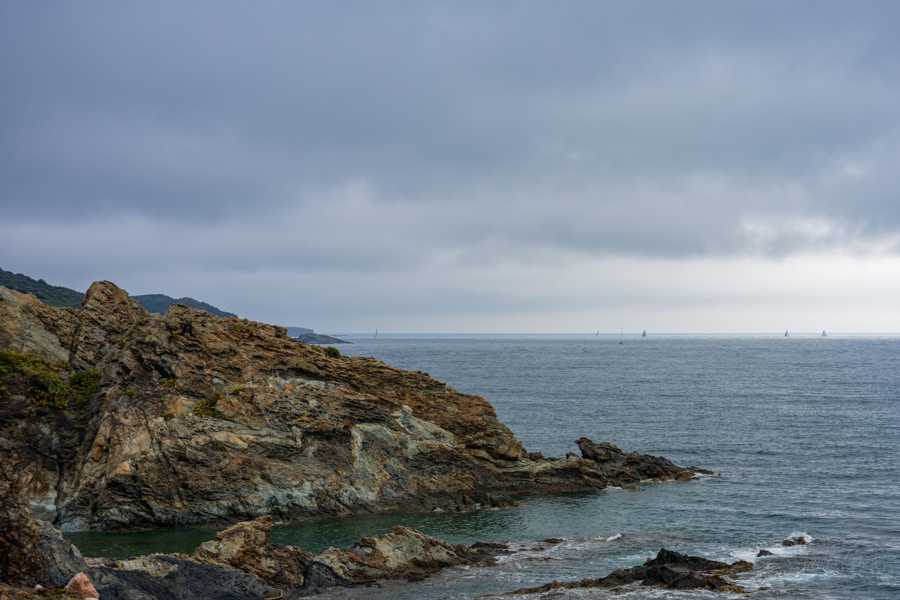 A rugged rocky headland of folded schist dropping into a green sea, distant sails on the horizon under a grey overcast sky.