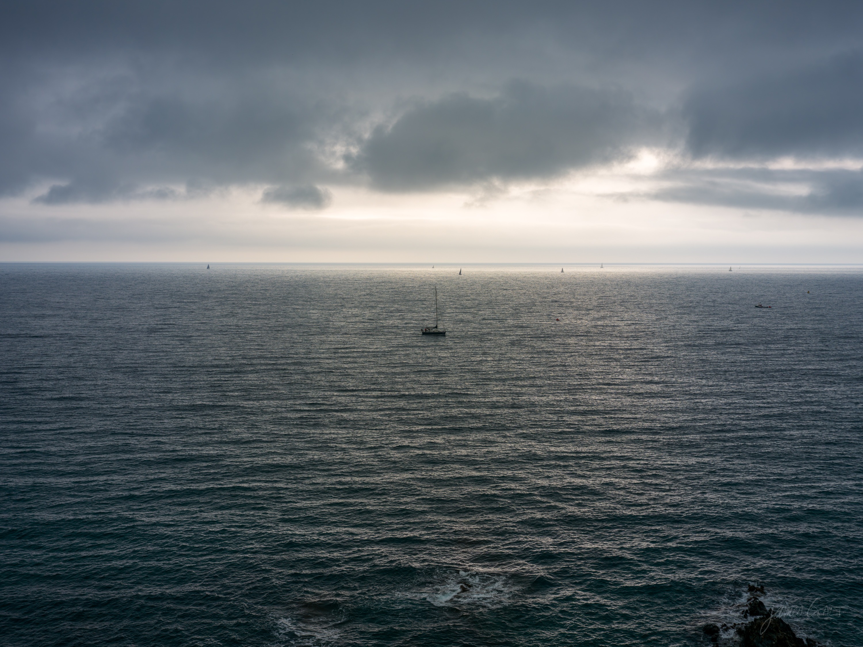 A wide flat grey sea under heavy cloud, a single yacht far out, a band of light along the horizon.