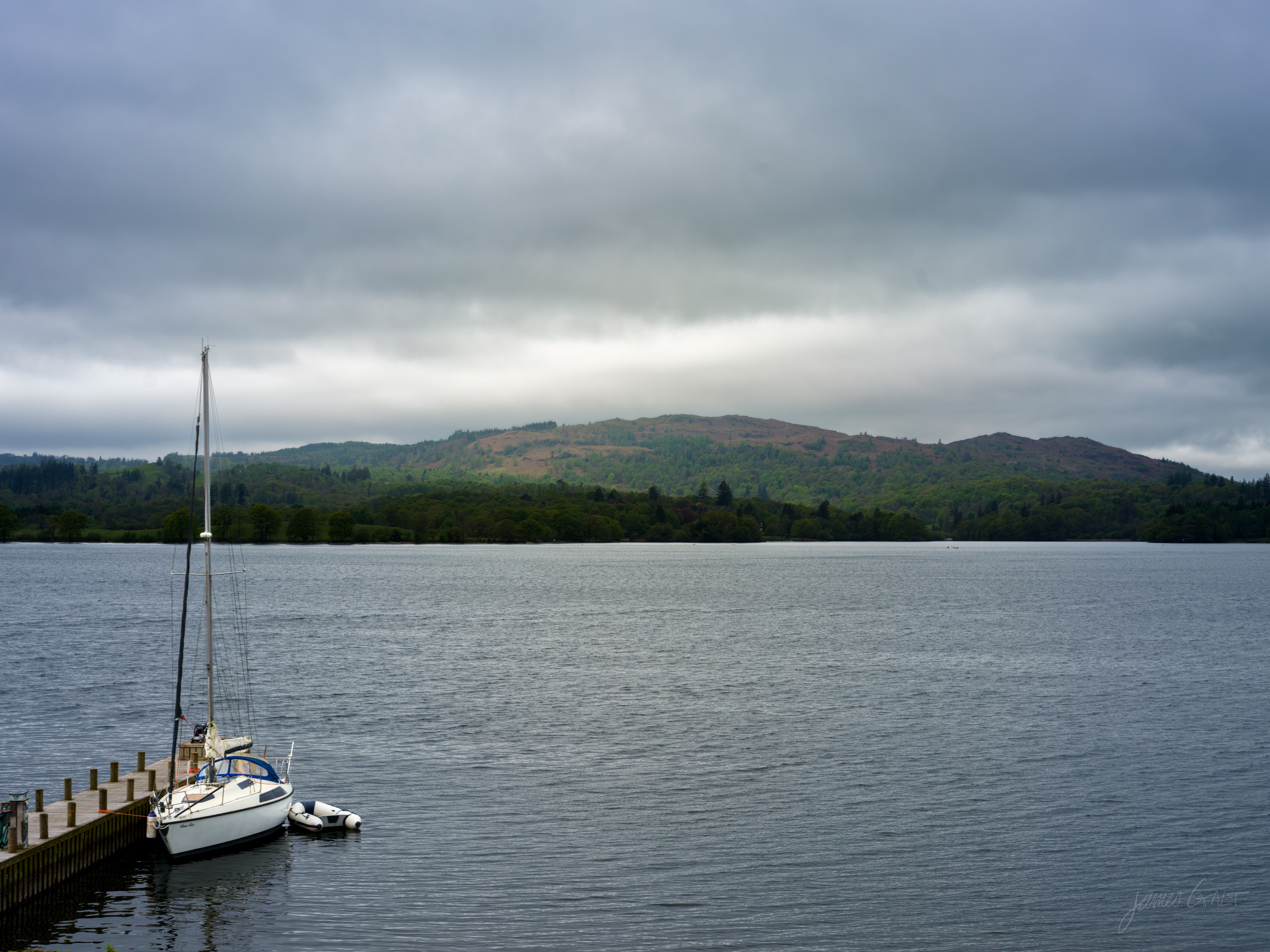 A single white yacht moored at a low jetty on a calm grey lake, wooded low hills opposite under a heavy overcast sky.