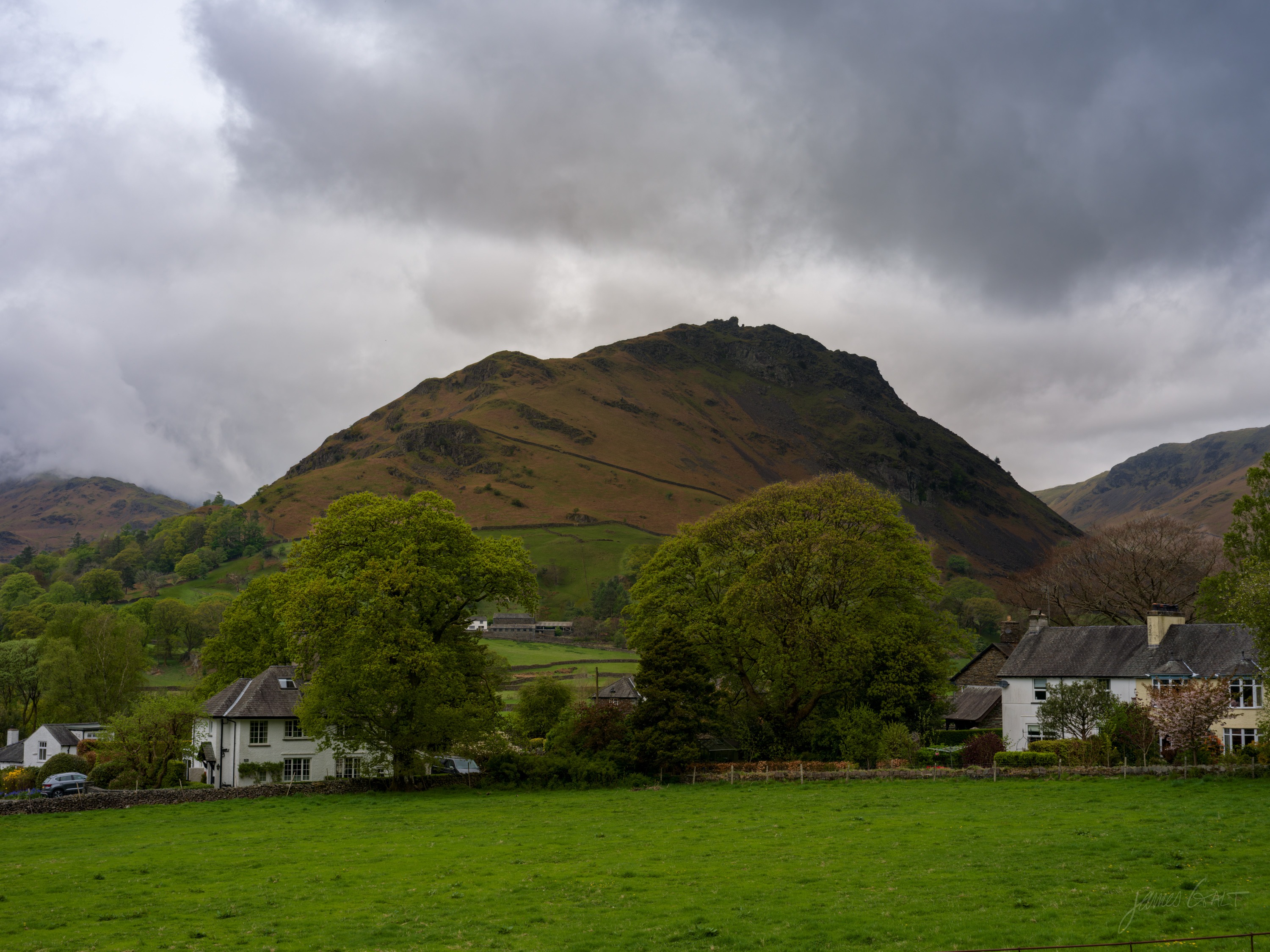 A steep pointed fell rising behind a cluster of white-rendered cottages and green trees, a green field in the foreground, cloud clinging to the higher ground.