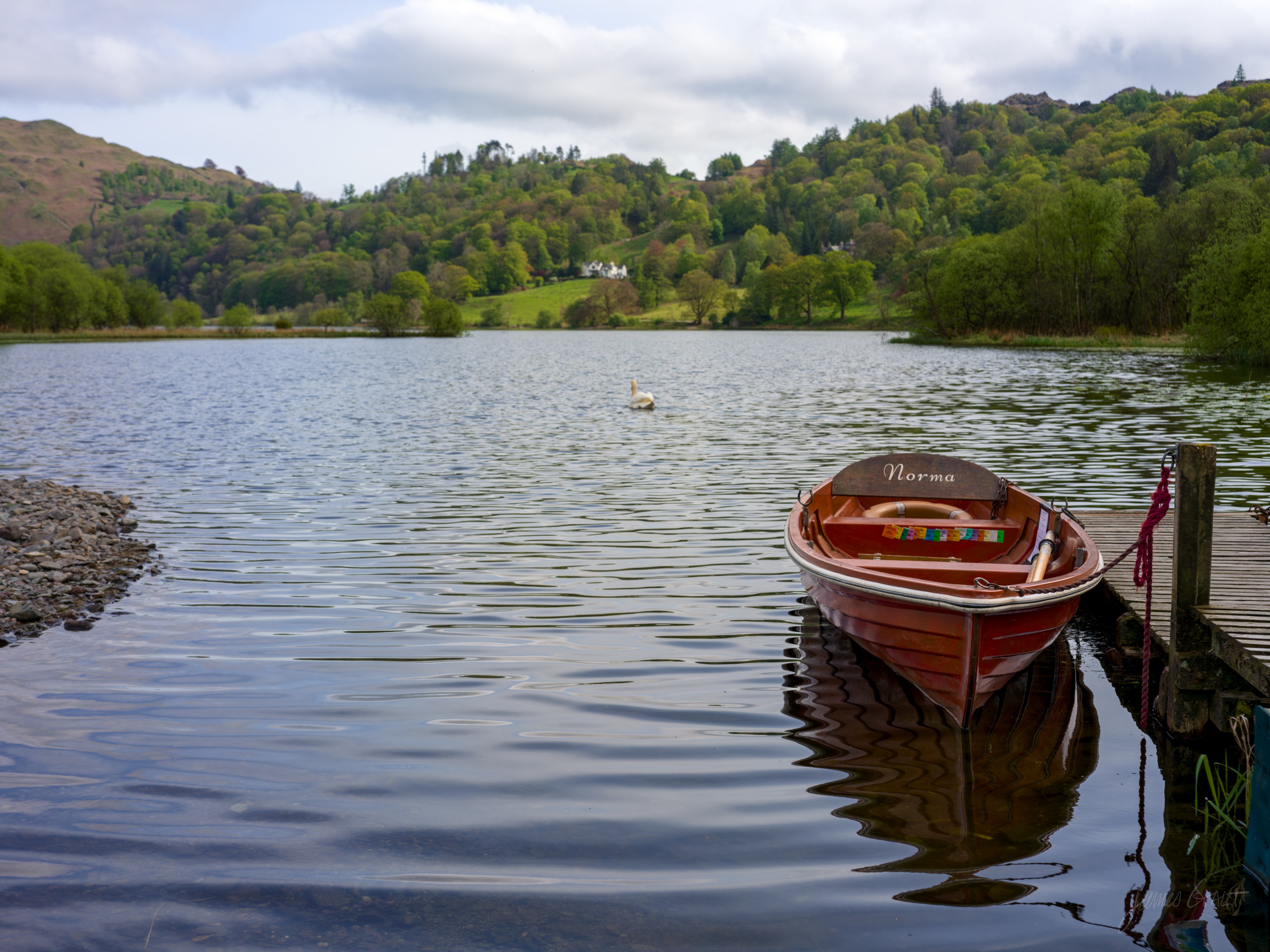 A red-and-varnish rowing boat named Norma moored at a small jetty on calm water, its reflection clear, a swan on the lake and wooded fells behind.