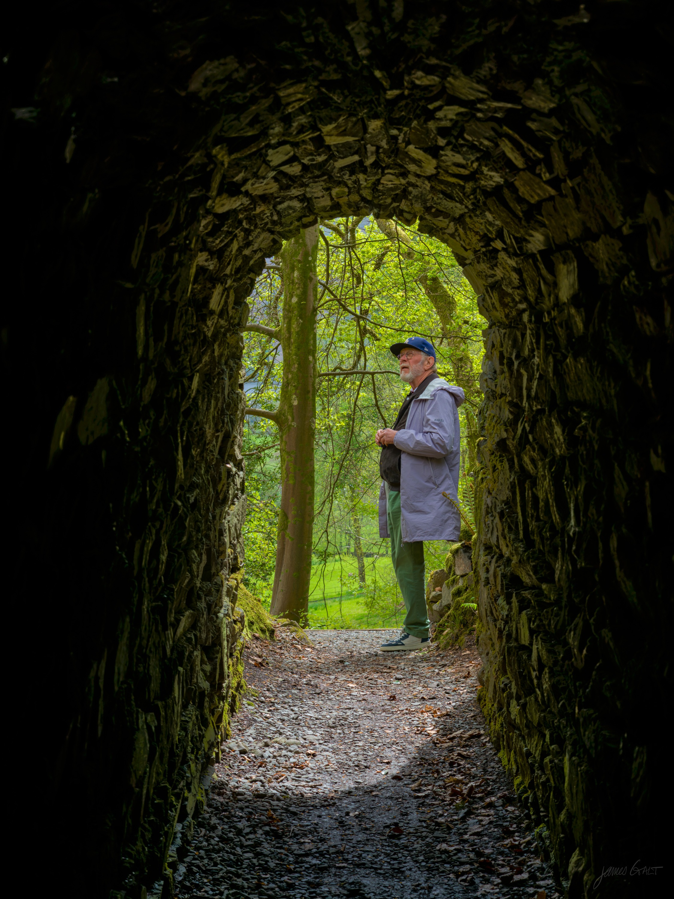 A man in a cap and grey coat standing inside a dark stone archway, framed against green woodland light beyond, rough stone around him.