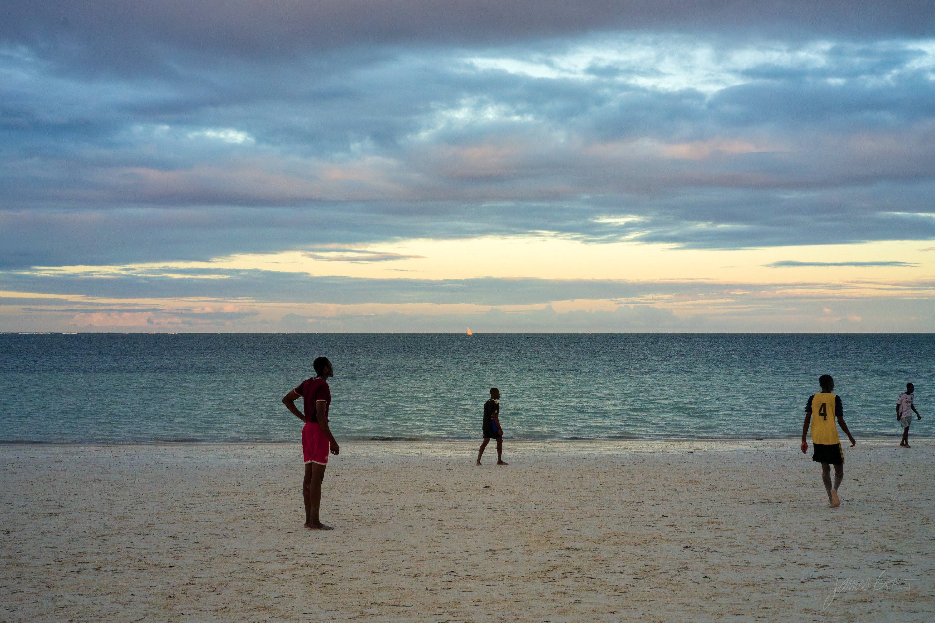 Figures scattered across a wide wet beach at dusk, their reflections in the film of water, a soft grey and gold sky above the sea.