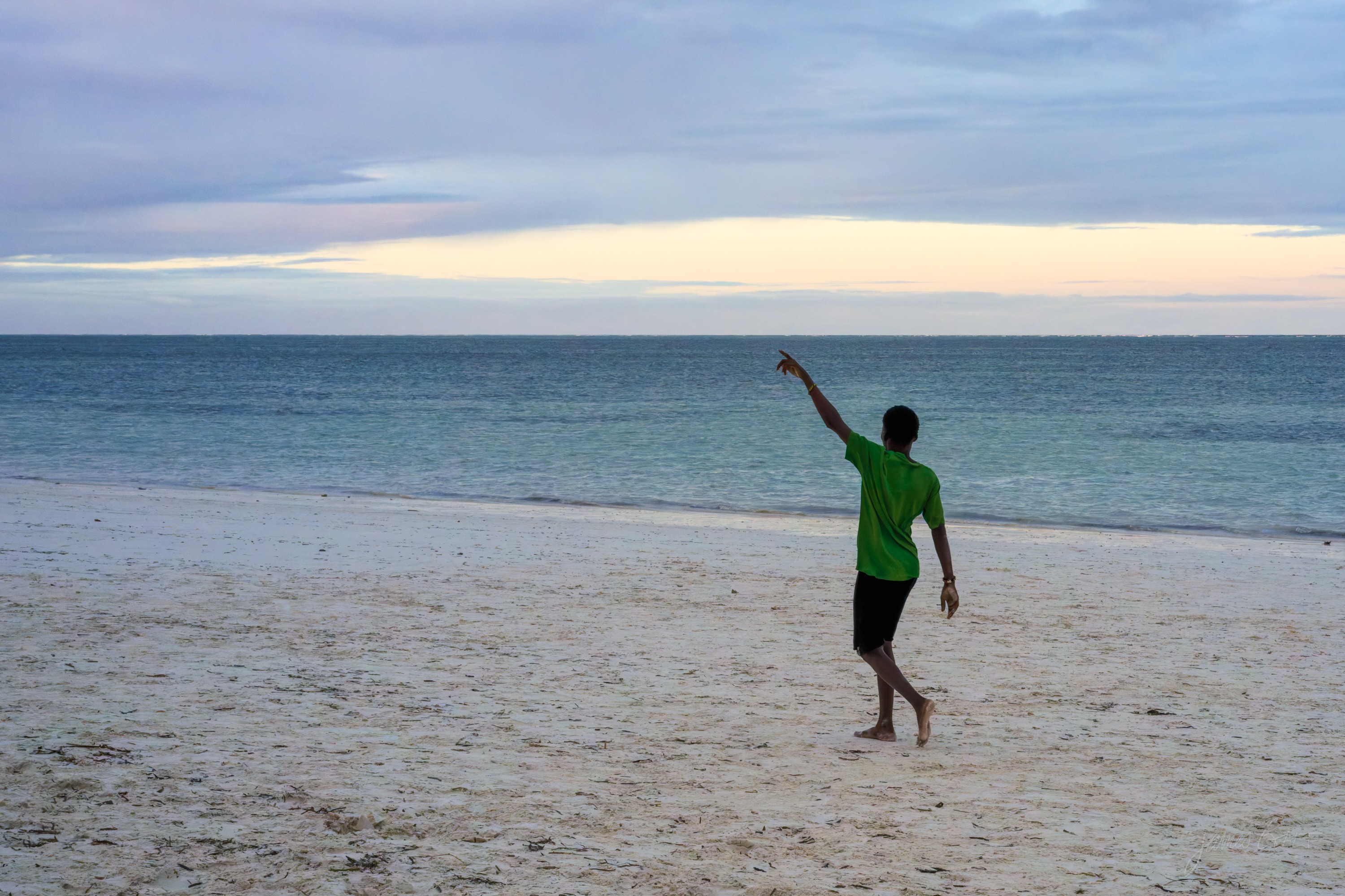 A person in a green shirt standing on the wet sand with one arm raised, waving something over his head, the sea and a pale sky behind.