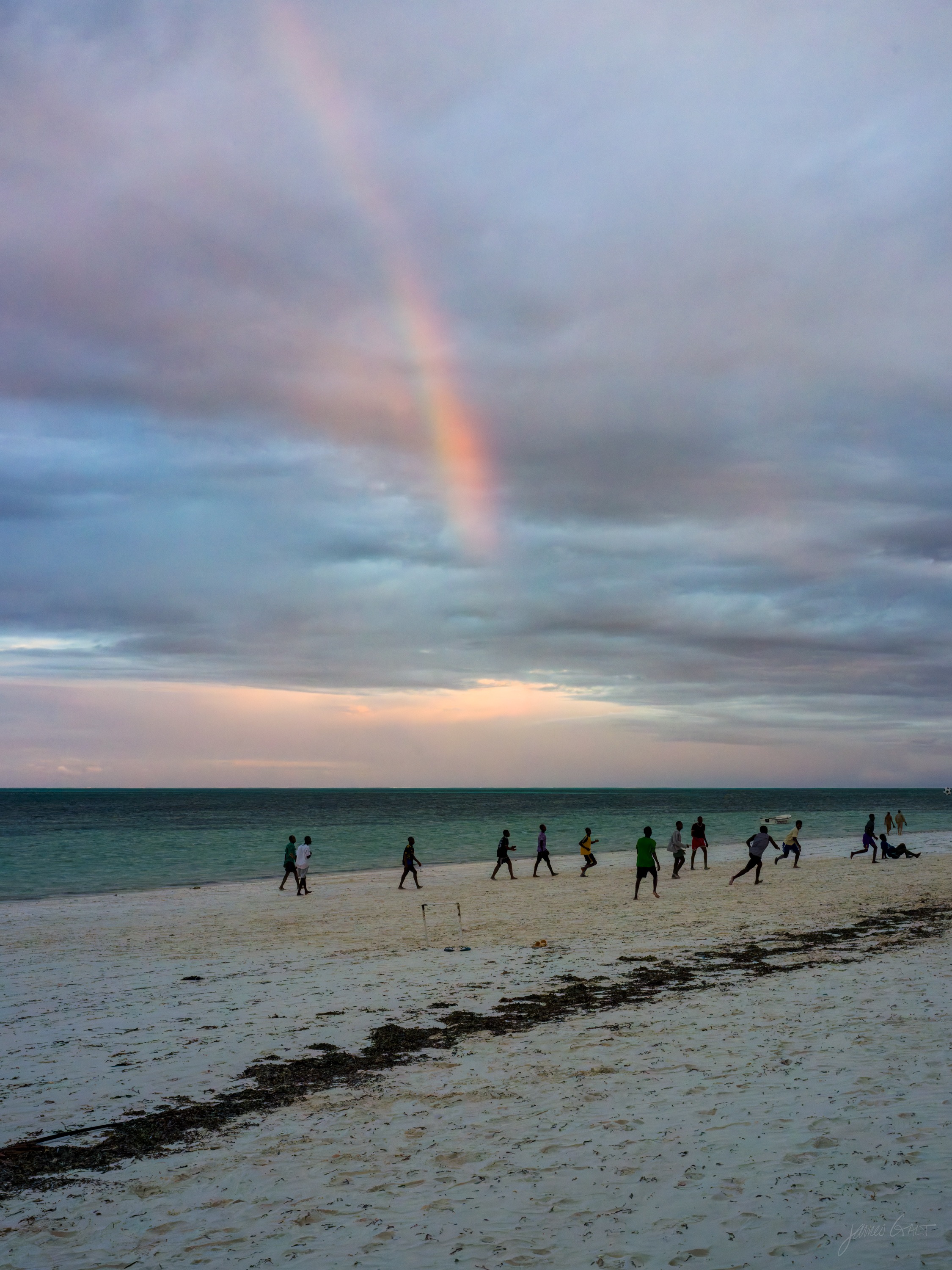 A faint full rainbow arching over a wide beach at dusk, small figures gathered on the sand beneath it, the sky heavy with cloud.