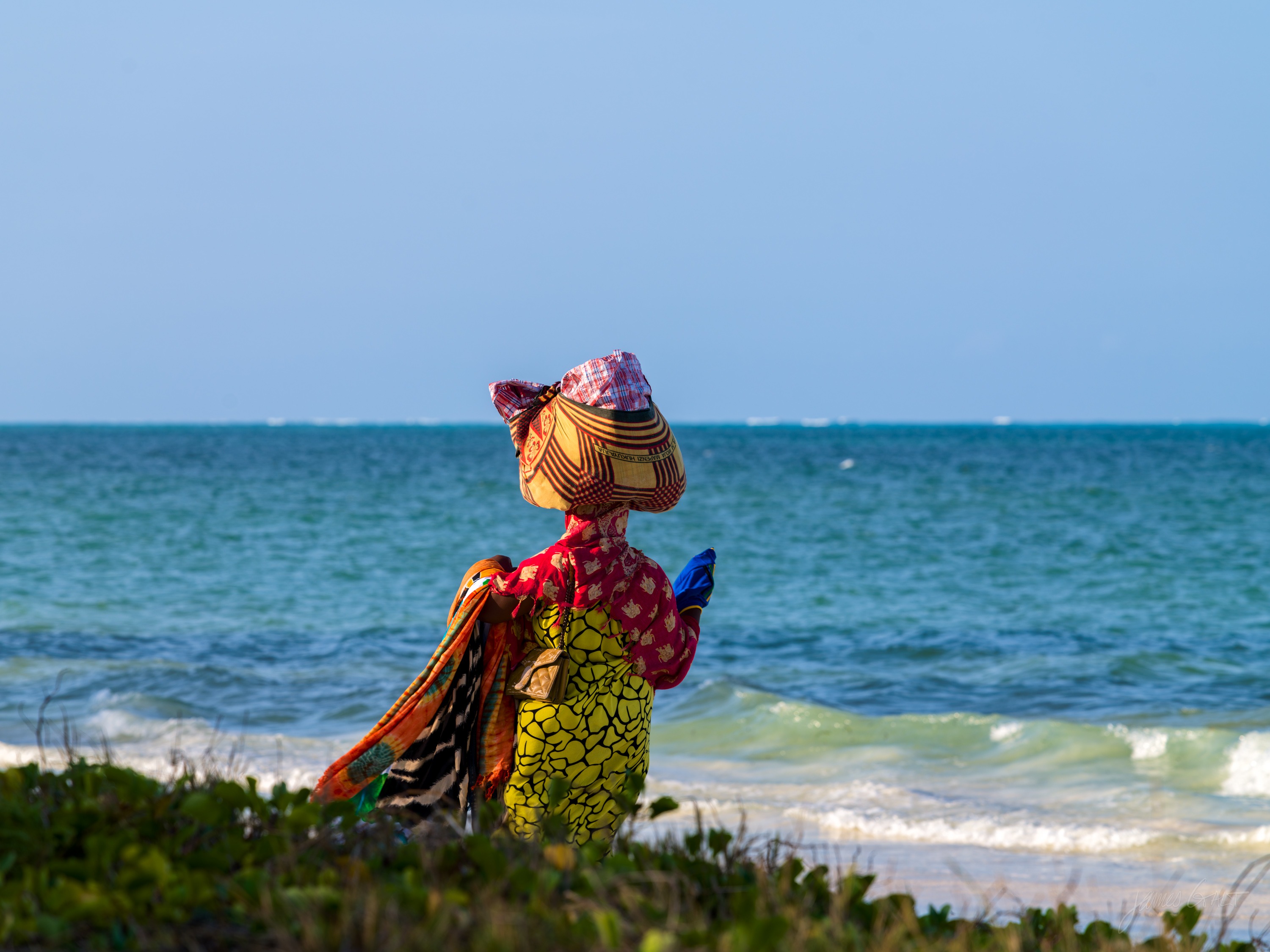 A woman in a bright, boldly patterned dress and headwrap standing on a low grassy dune looking out to the turquoise sea, seen from behind.
