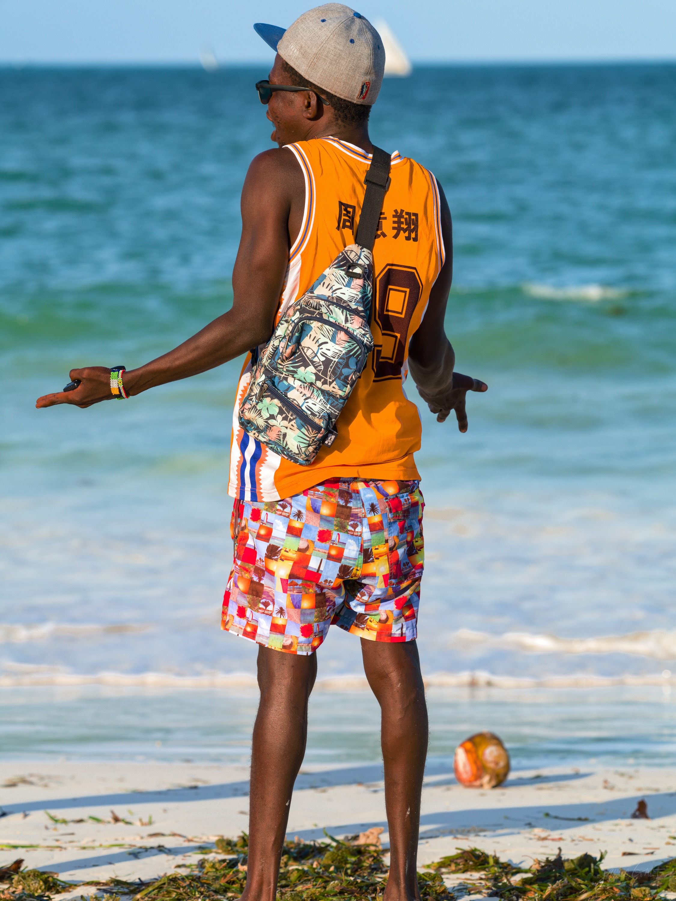 A man in an orange vest and patterned shorts standing on the beach facing the sea, a bag over his shoulder, turquoise water behind him.