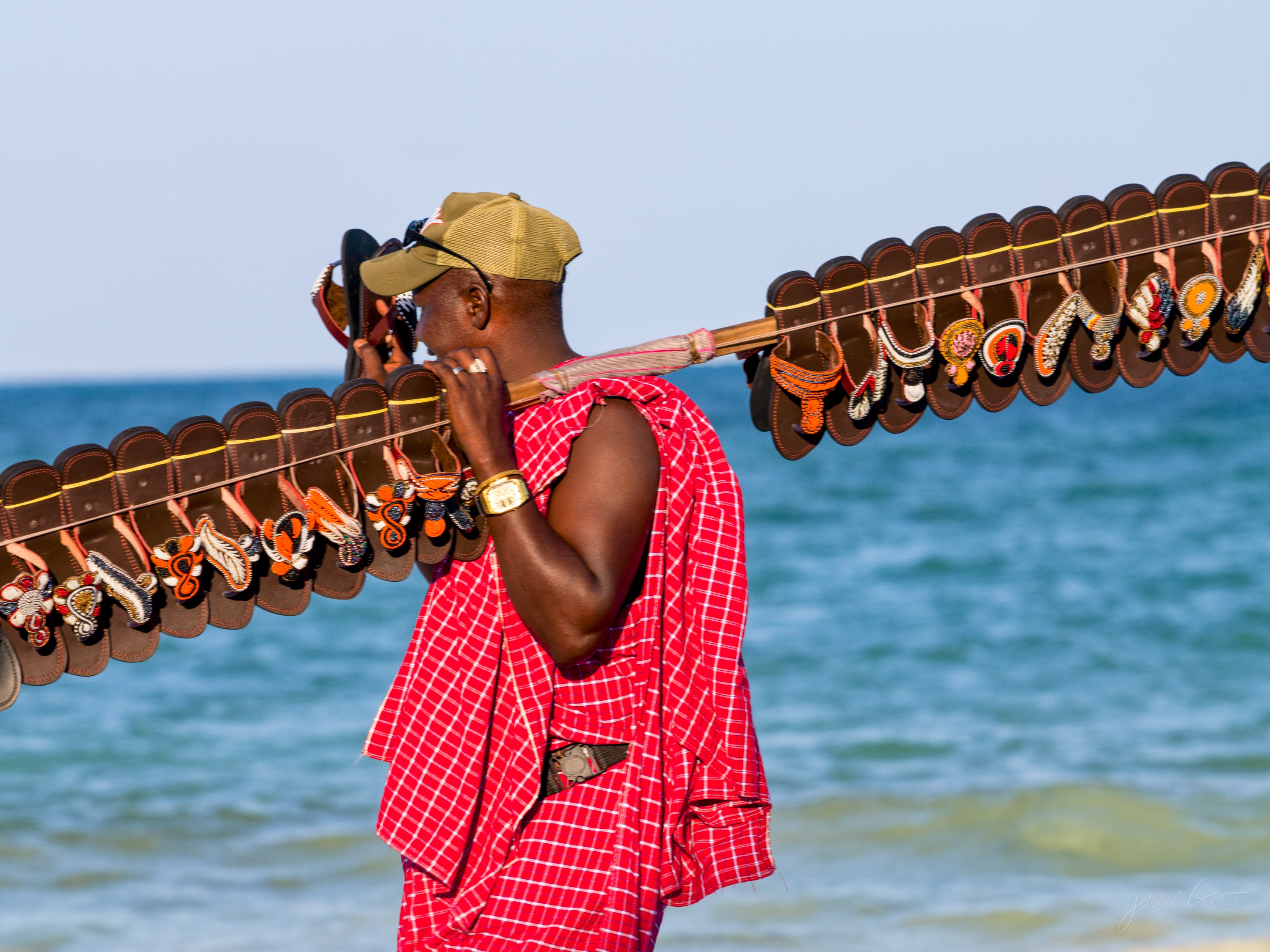 A man walking the beach carrying a long pole hung with carved souvenirs and necklaces over his shoulder, the sea behind him.
